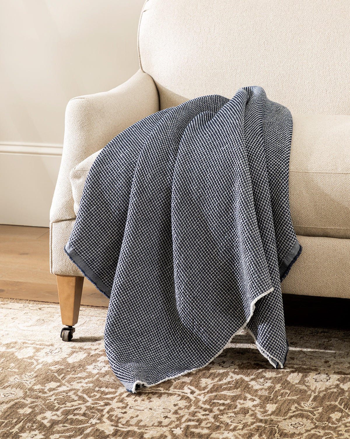 The Rebecca Throw, featuring a blue and white waffle weave texture, is draped over the arm of a light beige upholstered sofa beside a patterned brown rug on a wooden floor.