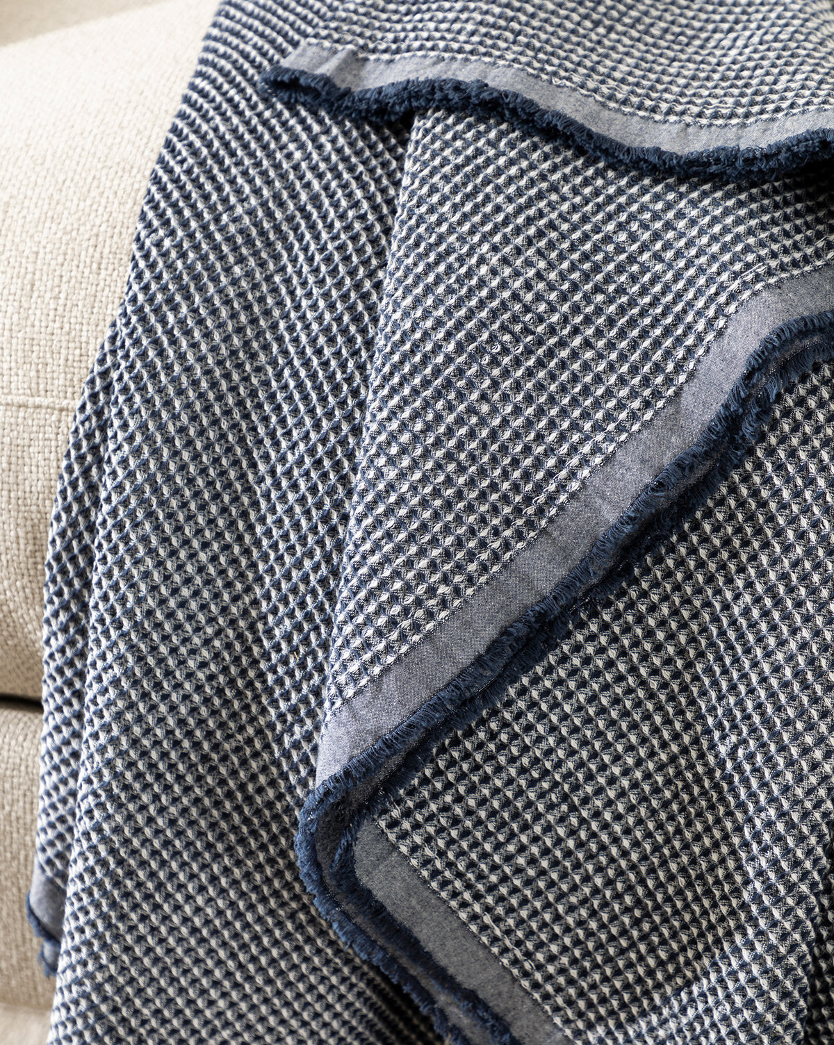 The Rebecca Throw, featuring a textured navy waffle weave and visible border, is draped over a light beige upholstered surface.