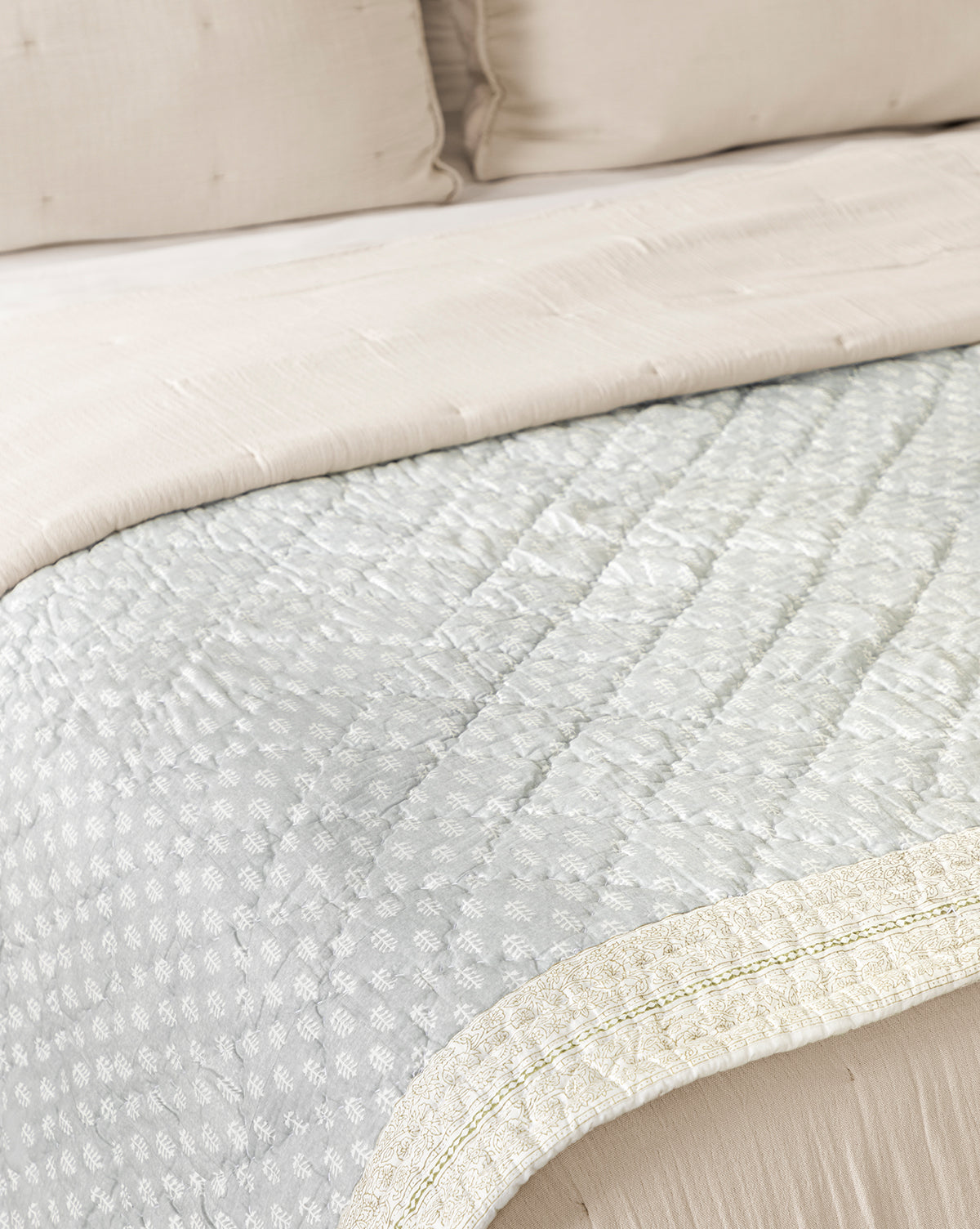 A close-up of a neatly made bed with cream-colored pillows and the Regatta Block Print Quilt, a handcrafted cotton piece topped with a light blue, patterned blanket featuring a decorative border by McGee & Co.