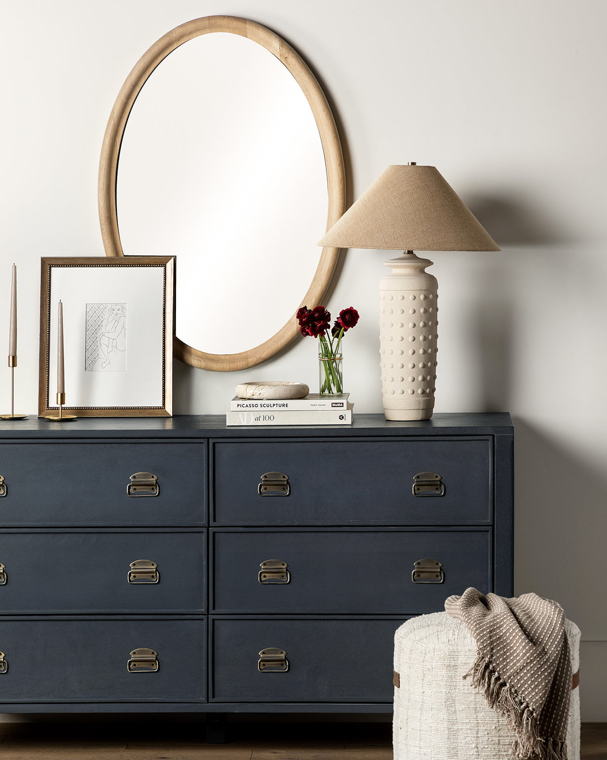 A navy blue Mackenna Dresser with brass pulls stands against a white wall, accessorized with a round wooden-framed mirror, lamp, books, framed art, candles, and red flowers. In the foreground sits a textured stool draped with a knit blanket.