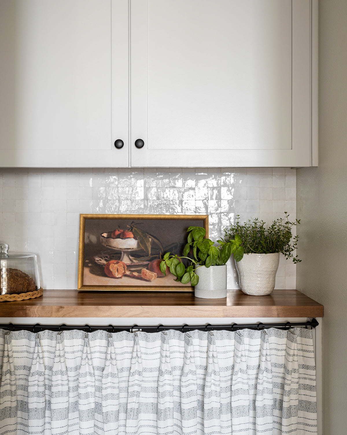 A cozy kitchen counter with potted basil and thyme, a ceramic bowl of mandarins, a glass jar of cookies, white cabinets above, lower cabinets hidden by a patterned curtain, and “Manderine Still Life by Pascal Giroud” framed on the wall.