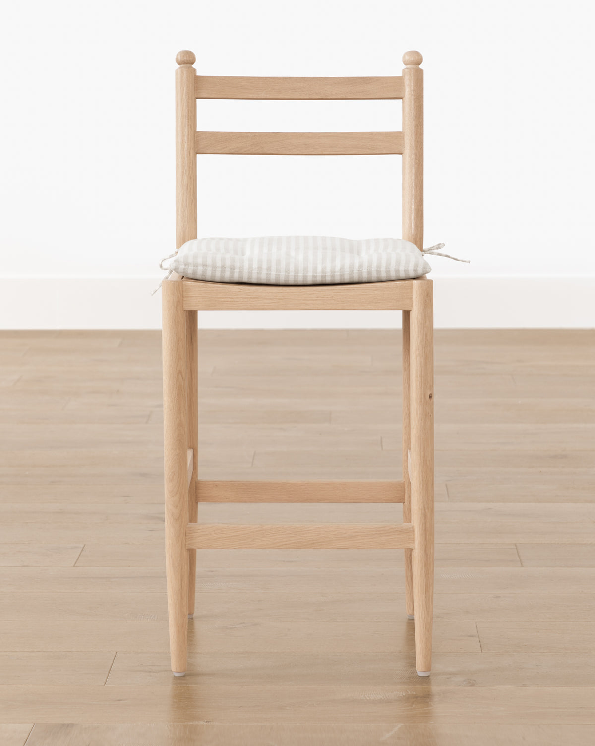 The Romany Counter Stool, with straight wooden legs and a ladder backrest, features a light checkered seat cushion tied to the seat and sits on a light wood floor against a plain white wall, showcasing modern simplicity and style, by McGee & Co.