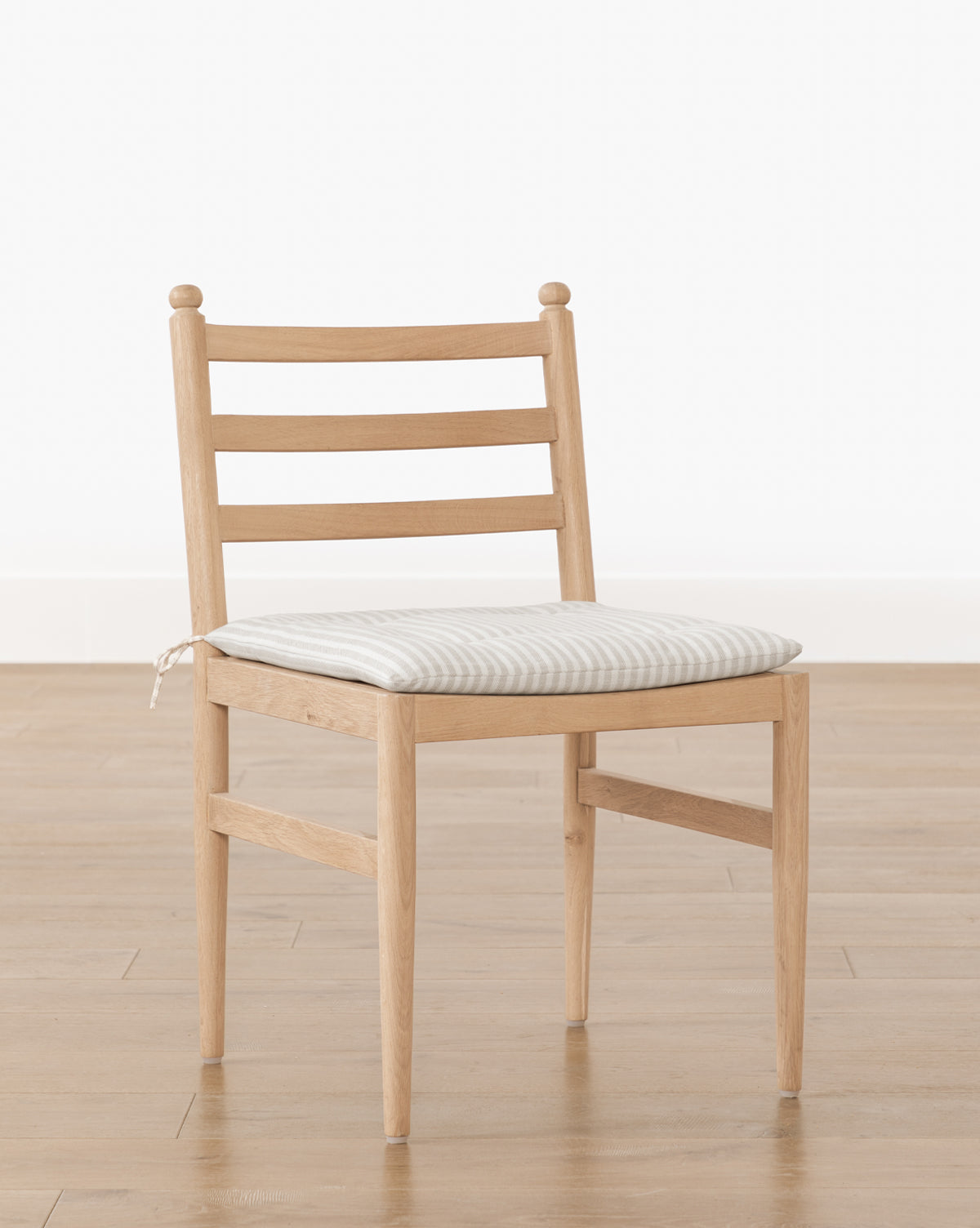The Romany Dining Chair features a simple white oak frame, ladder-back design, and a light linen seat cushion, shown on a wooden floor against a plain white background by McGee & Co.