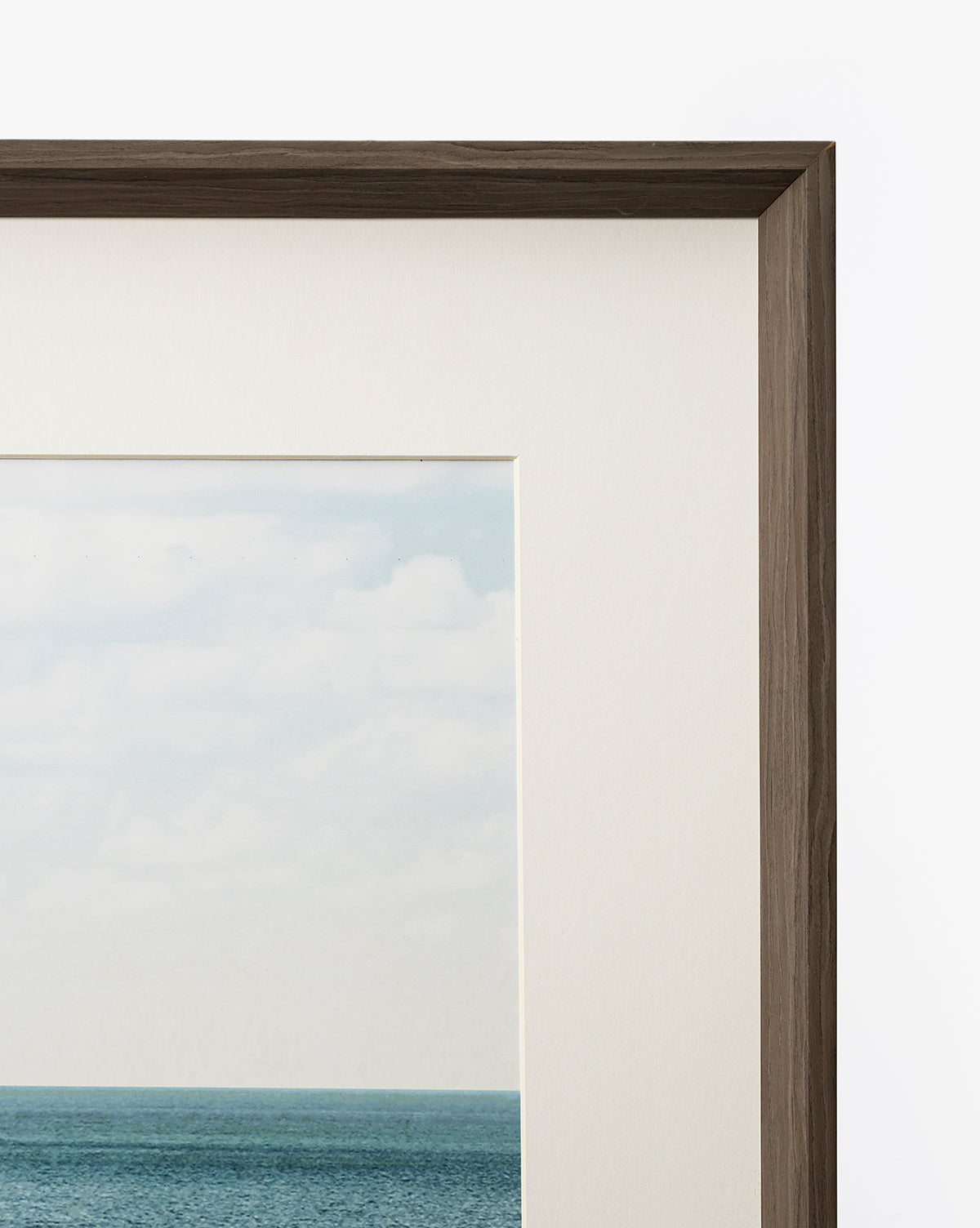 A close-up of the top right corner of Room with a View, a gallery-quality art piece showing a cloudy sky, blue ocean, off-white mat, and dark walnut frame set against a white background by McGee & Co.