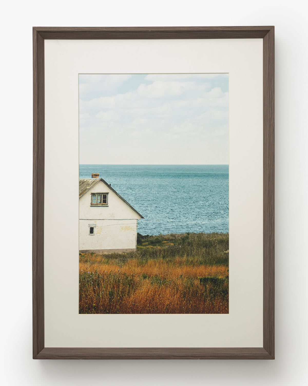 Room with a View: A gallery-quality framed photograph of a white house by the sea, tall grasses in front, and a calm blue ocean under cloudy skies, all set in a dark walnut frame, by McGee & Co.