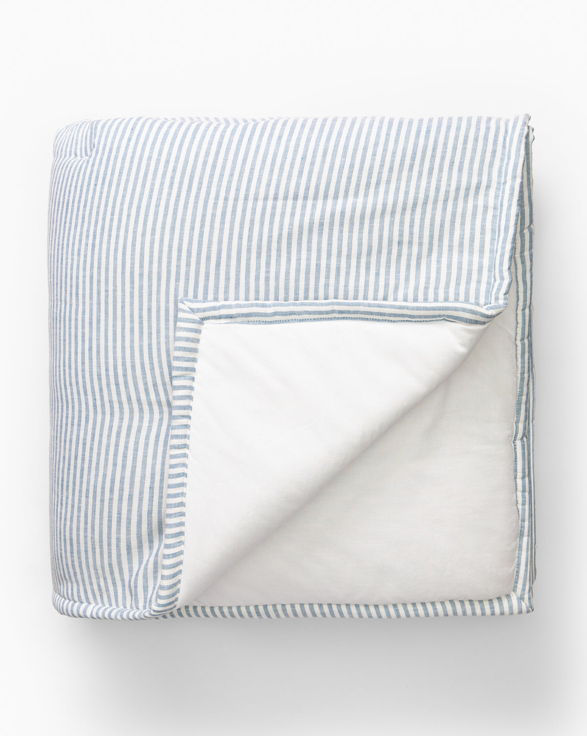 The Rosemary Quilt, shown folded on a white background, features blue and white vertical stripes on one side and solid white on the reverse in yarn-dyed linen-cotton. A turned corner reveals the contrasting side.