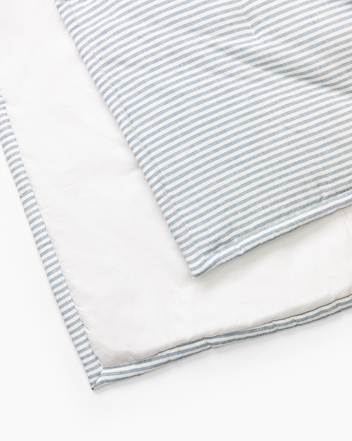 The Rosemary Quilt is folded to reveal its blue and white striped front and plain white back. Crafted from soft linen and cotton, this yarn-dyed quilt adds cozy style when displayed on a white surface.