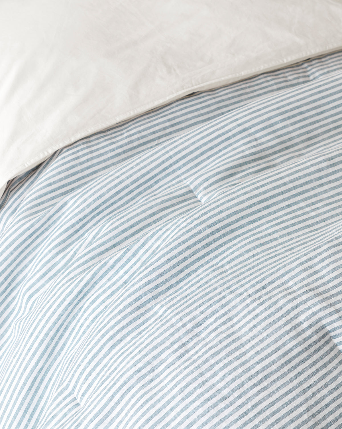 A close-up of a bed features the Rosemary Quilt, displaying soft, light blue and white striped linen-cotton fabric with a white pillow. The neatly arranged bedding creates a tidy and inviting look.