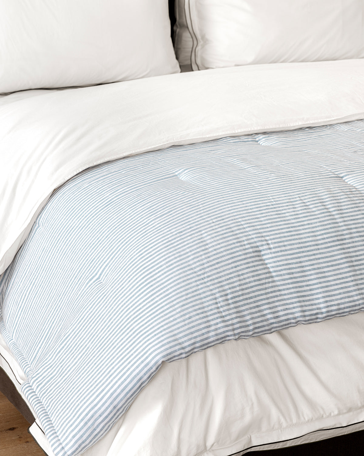 A neatly made bed features soft linen and cotton bedding with white pillows, a white duvet, and the Rosemary Quilt in light blue and white stripes folded at the foot for a serene look.