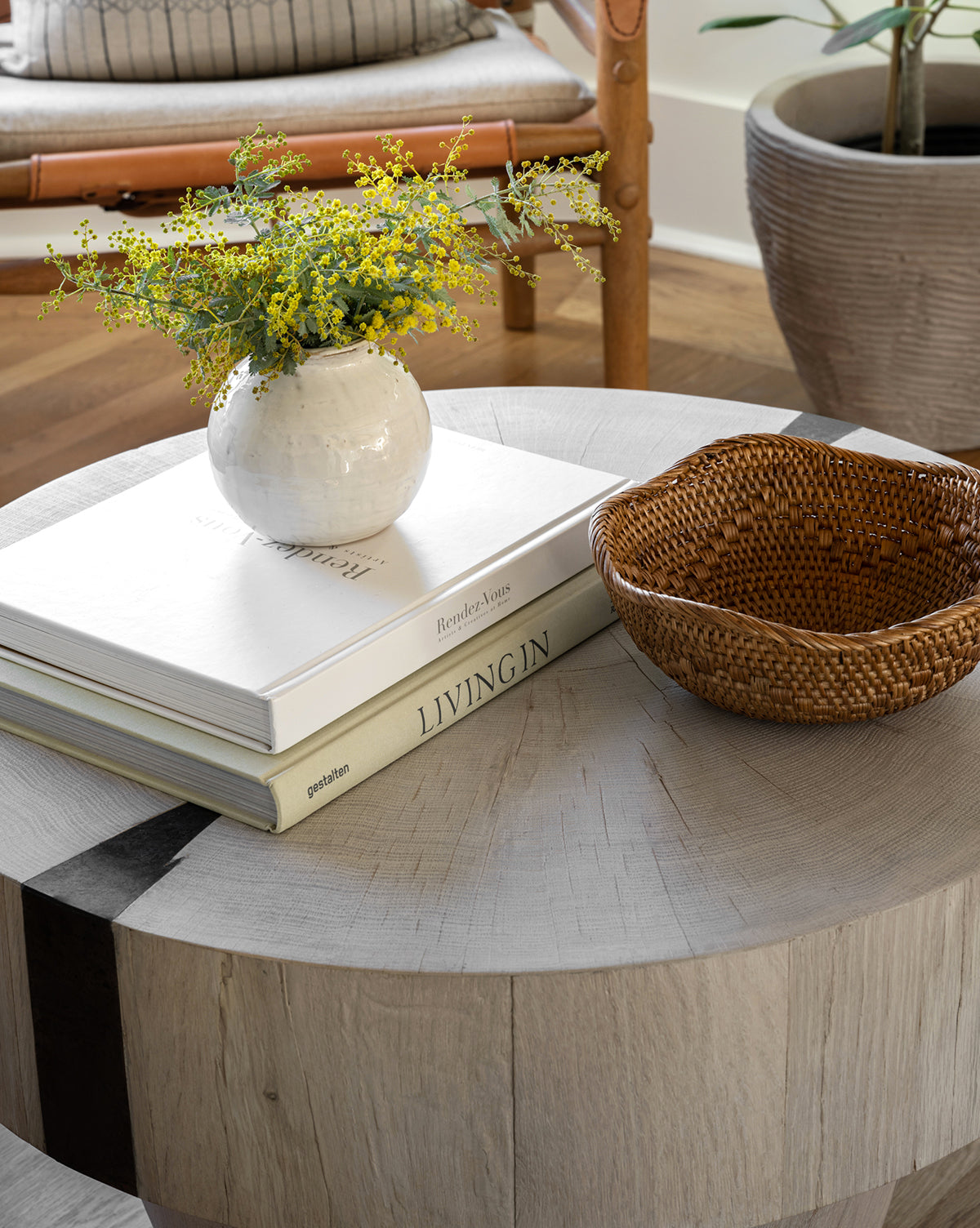 A round wooden coffee table displays two stacked books, the Rounded Ceramic Vase with yellow flowers, and a woven basket for inviting charm. In the background, a potted plant and a chair complete your cozy home decor.
