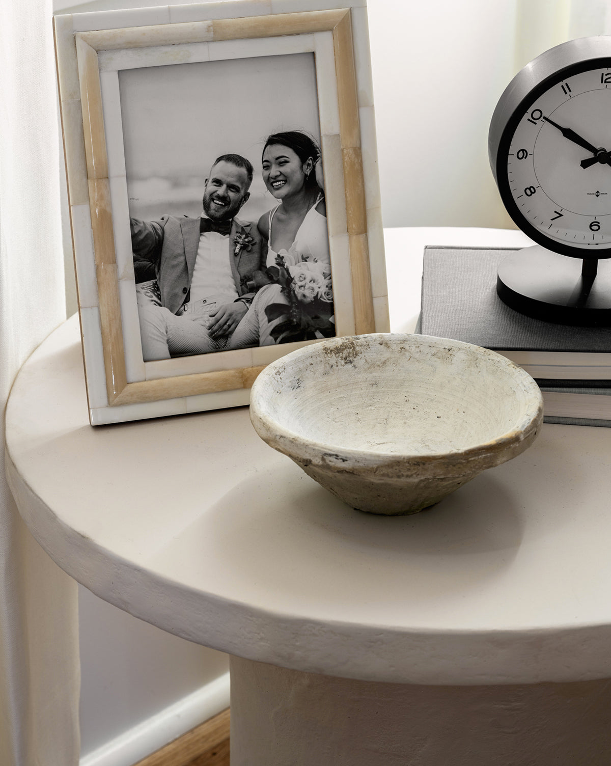 A round engineered wood table holds a ceramic bowl, books, a clock set to 9:10, and a black-and-white wedding photo in an Inlay Bone Frame beside a window with soft lighting.