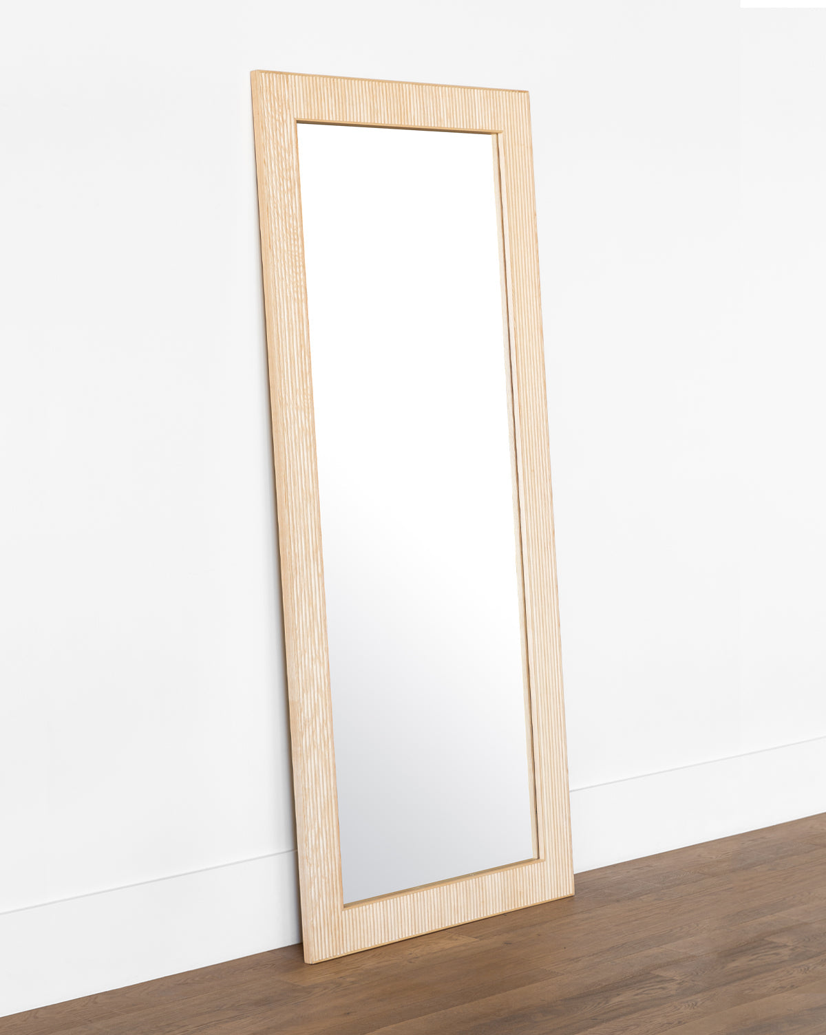 The Rowan Floor Mirror is a large rectangular mirror with a ribbed oak frame, leaning against a white wall on a brown wooden floor by McGee & Co.