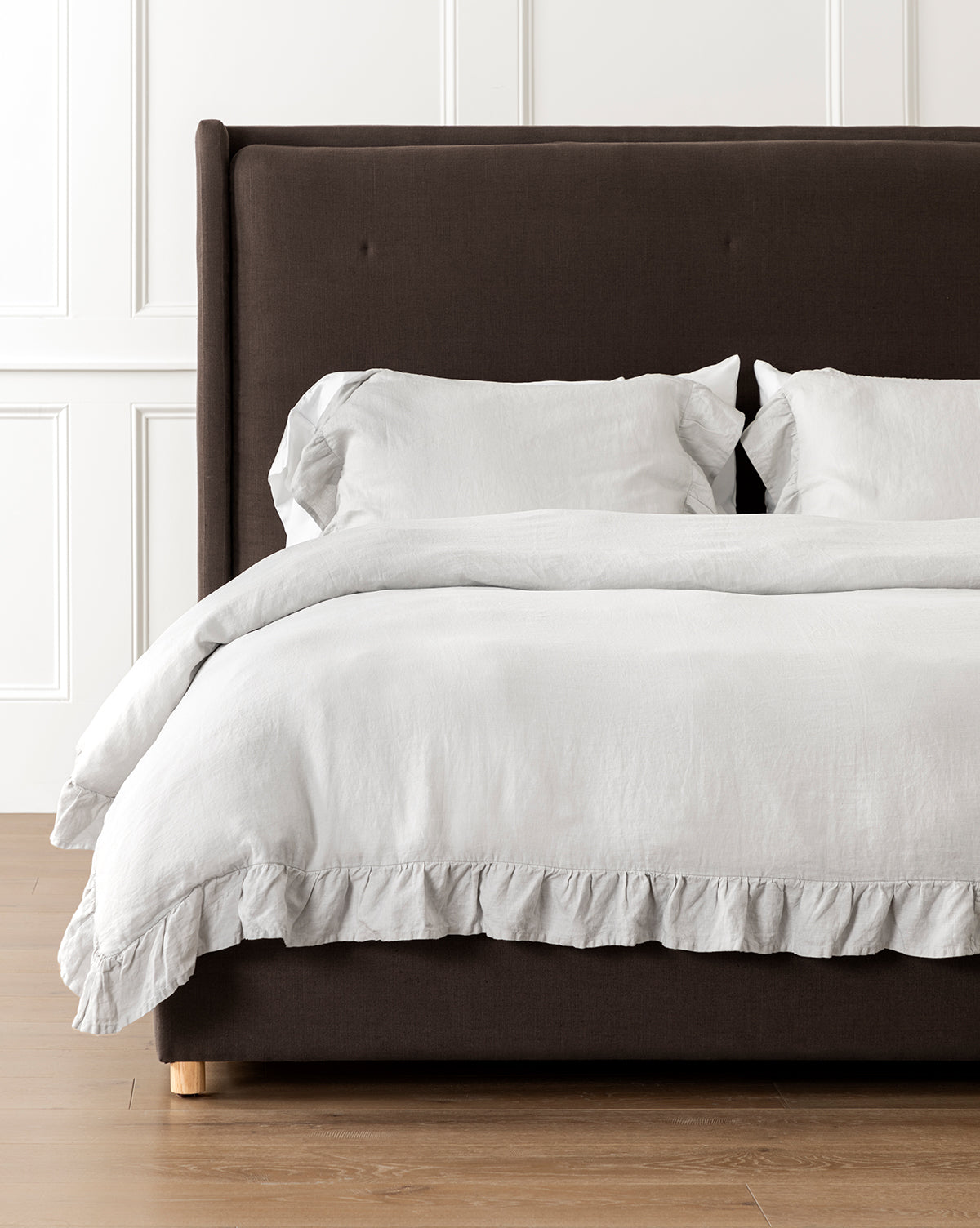 A neatly made bed featuring the Ruffle Linen Duvet Cover and two pillows, set against a dark upholstered headboard and white paneled wall on a light wood floor.