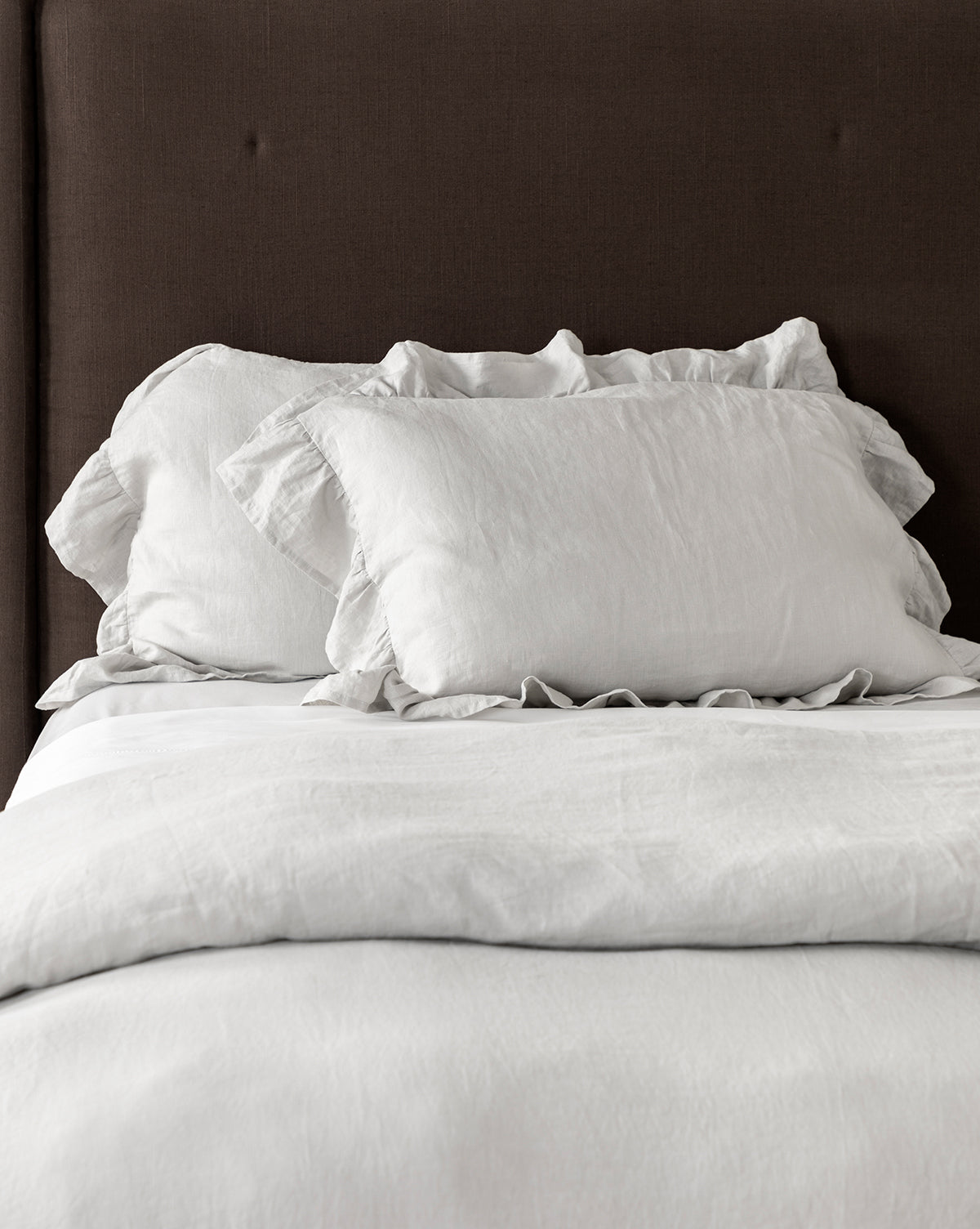 A neatly made bed with a dark brown upholstered headboard features romantic white bedding, including ruffled-edge pillows highlighted by Ruffle Linen Shams (Set of 2), plus a matching white comforter and sheets.