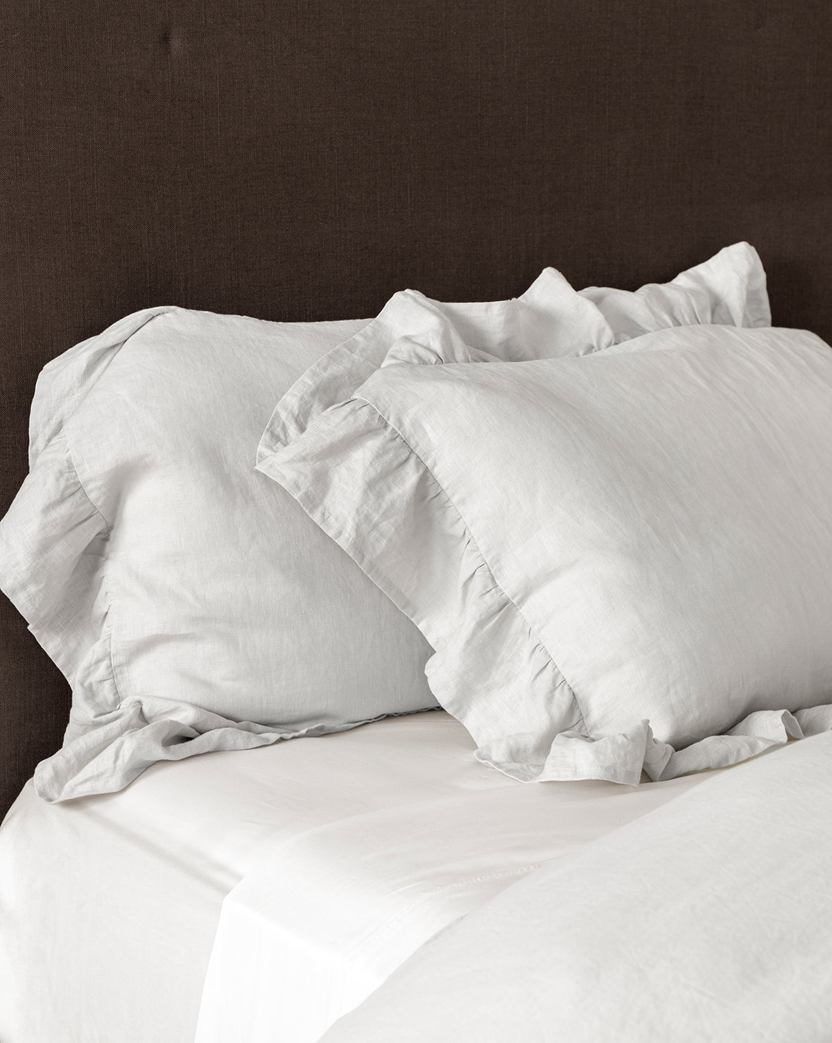The Ruffle Linen Shams (Set of 2) add a romantic accent to a neatly made bed with white sheets, complemented by a dark brown upholstered headboard.