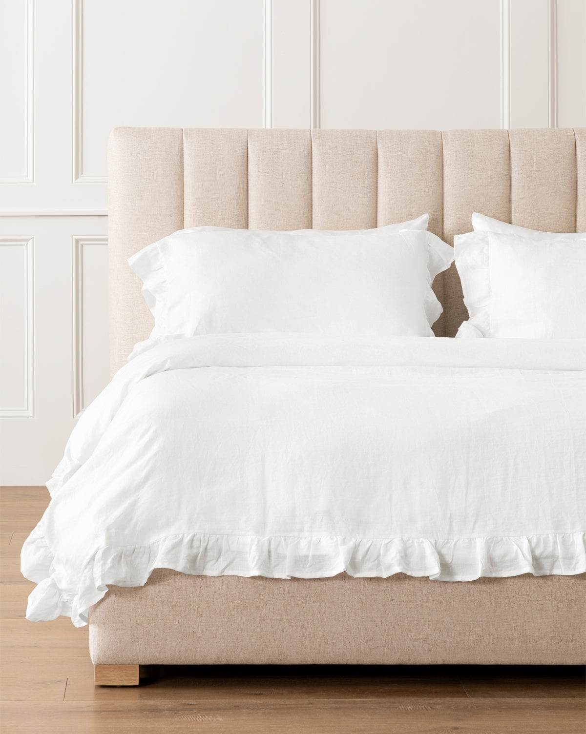 A bed with a beige upholstered headboard is dressed in the Ruffle White Linen Duvet Cover and matching pillows, set against a white paneled wall on a wooden floor.