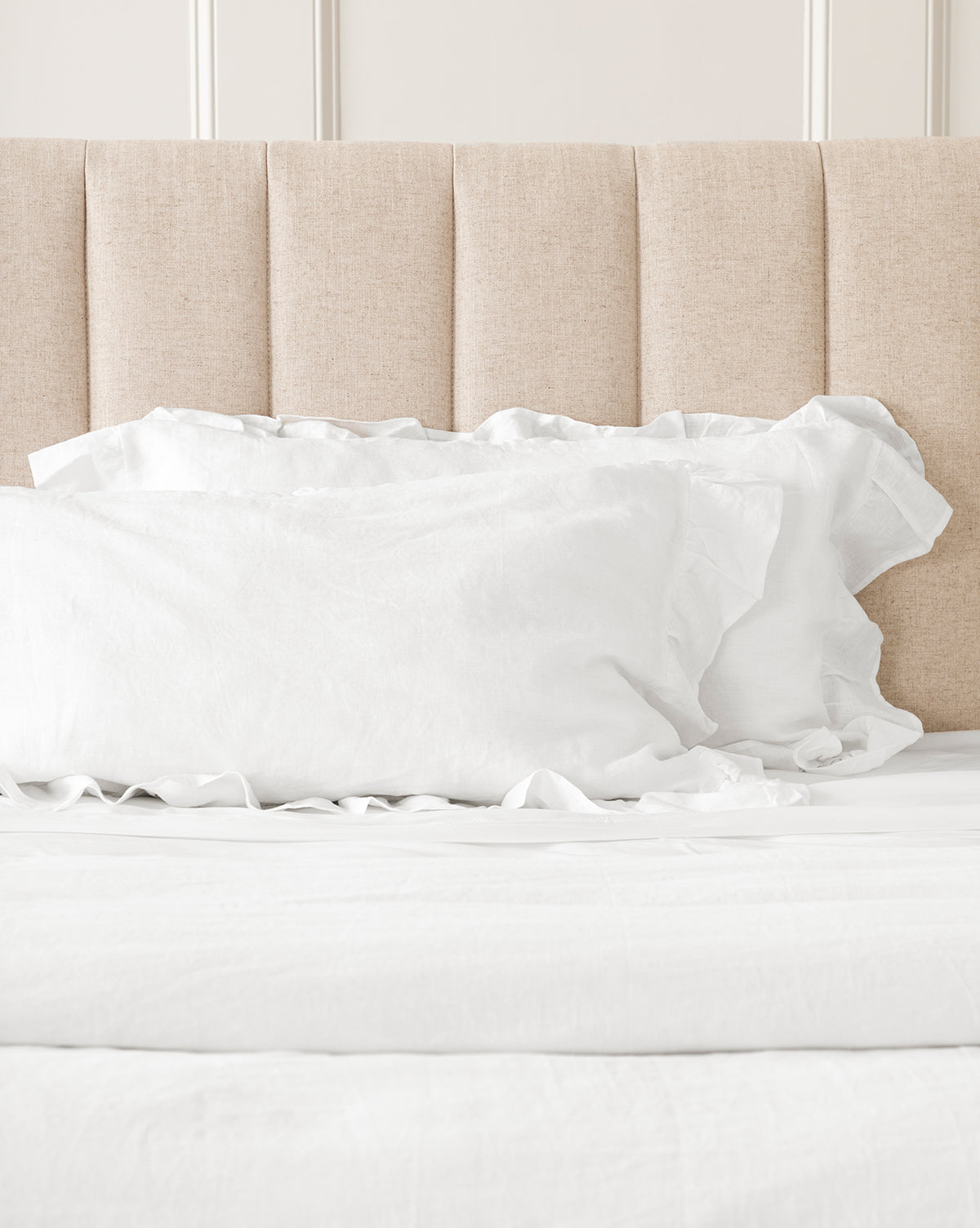 A bright, minimalistic room features a neatly made bed with a beige upholstered headboard and Ruffle White Linen Shams (Set of 2), all crafted from 100% linen.