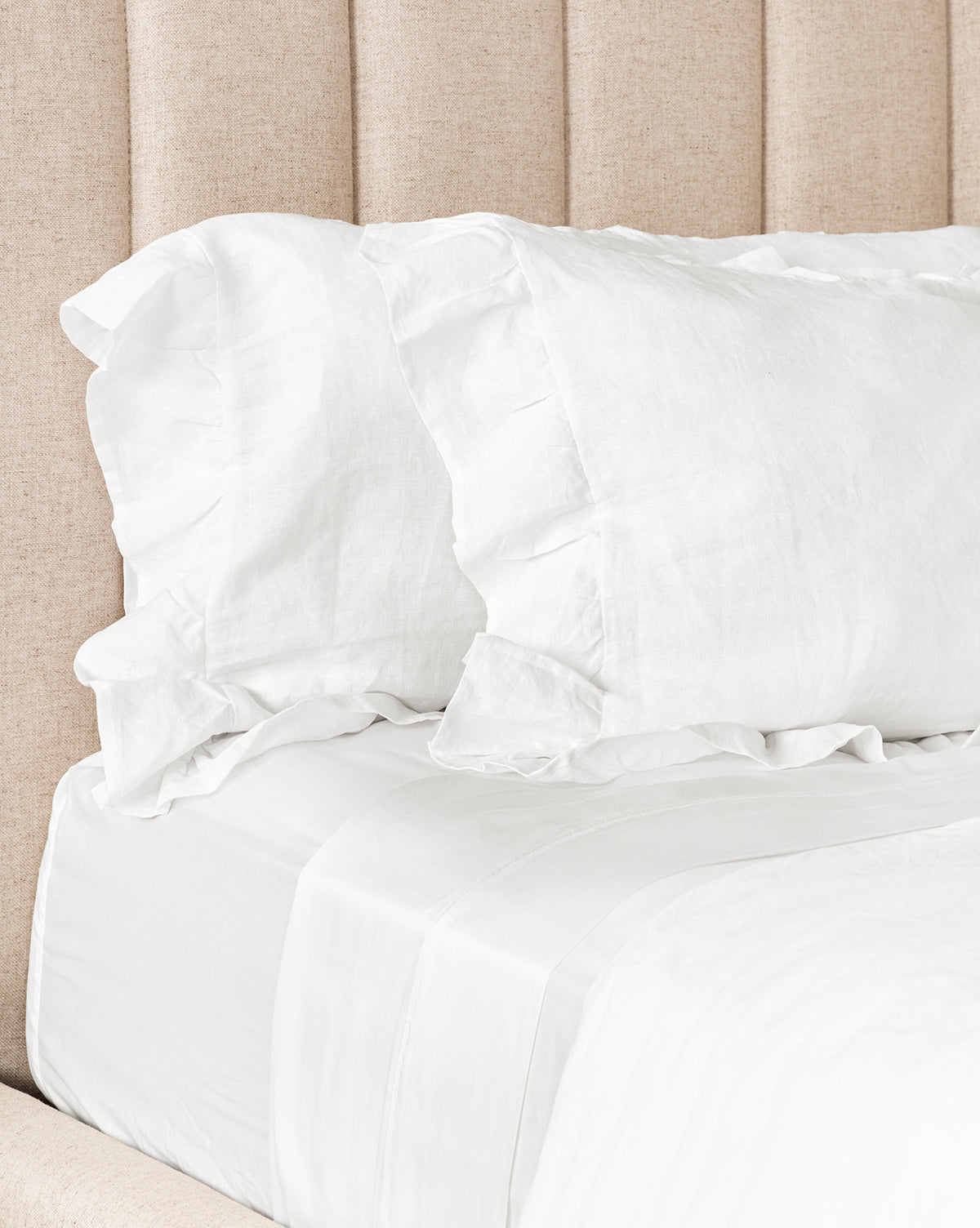 A neatly made bed with crisp white sheets and Ruffle White Linen Shams (Set of 2), featuring ruffle details, is set against a beige upholstered headboard with vertical panels.