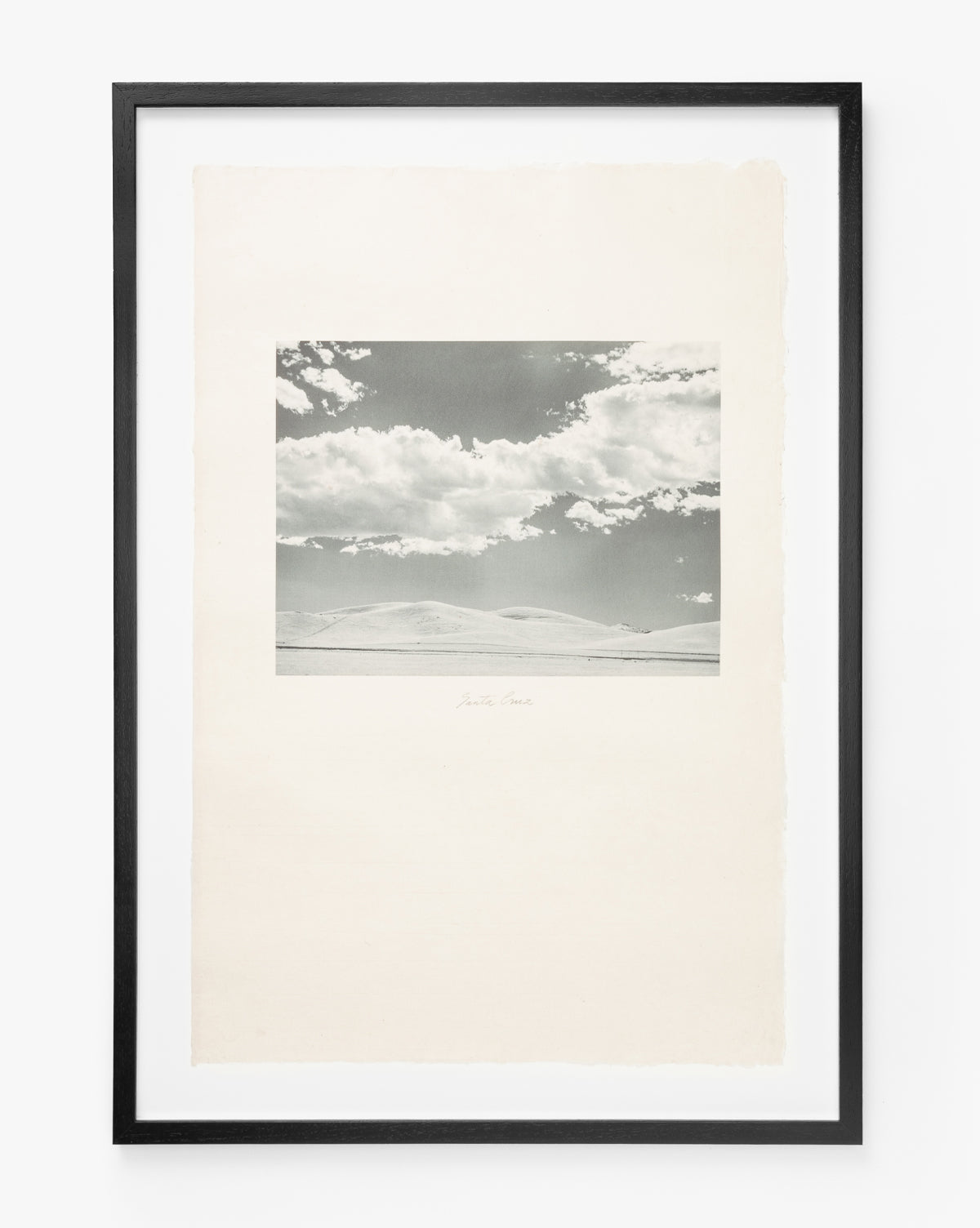 Black-and-white Santa Cruz photo in a black frame captures a sweeping coastal view with rolling hills and clouded skies. Centered on a white mat with rough edges and handwritten text, it creates timeless vintage wall art.