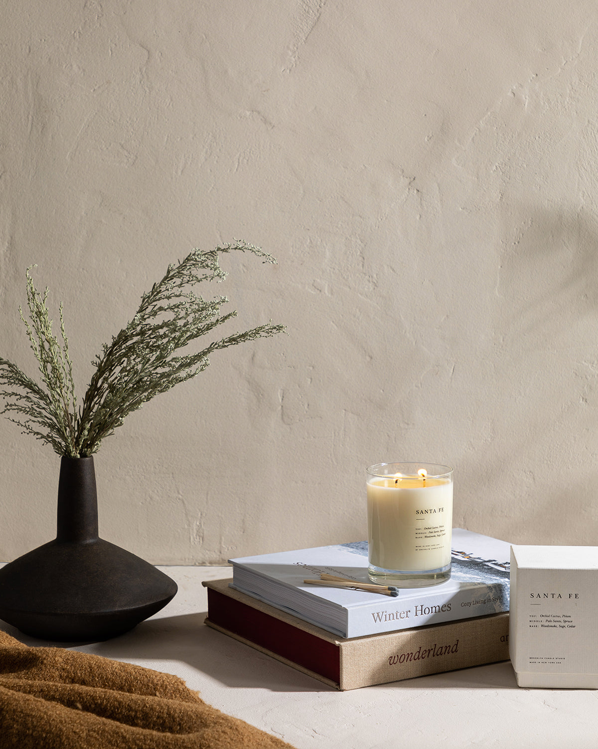 A dark vase with green sprigs, stacked books, a lit Santa Fe Candle in a glass jar, and its box rest on a beige surface against a textured wall, with brown fabric and touches of palo santo in front.