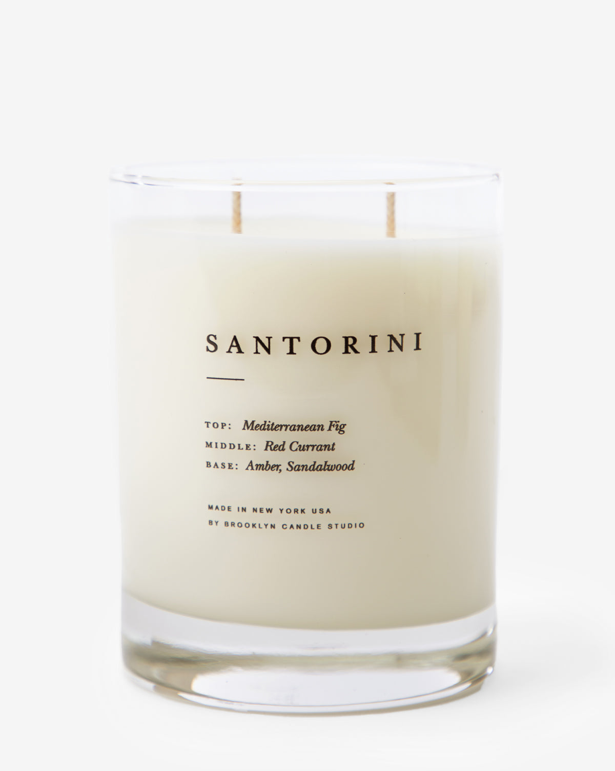 The Santorini Candle is a white soy wax candle in a clear glass jar labeled SANTORINI, featuring Mediterranean Fig, Red Currant, Amber, and Sandalwood notes with two wicks and a minimalist design.