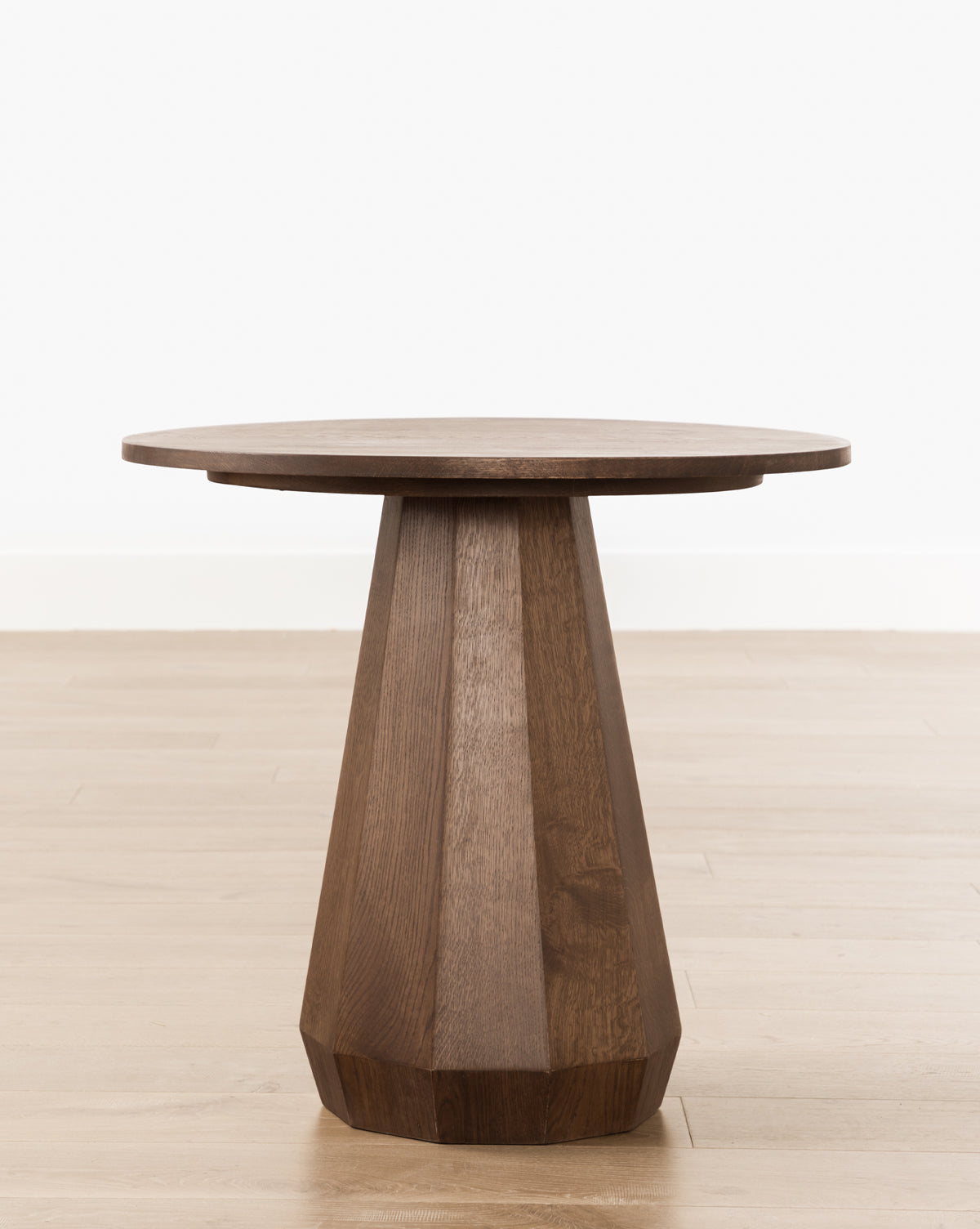 The Santos Side Table is a small, round table made from solid oak with a faceted geometric pedestal base, shown on a light wood floor against a plain white wall by McGee & Co.