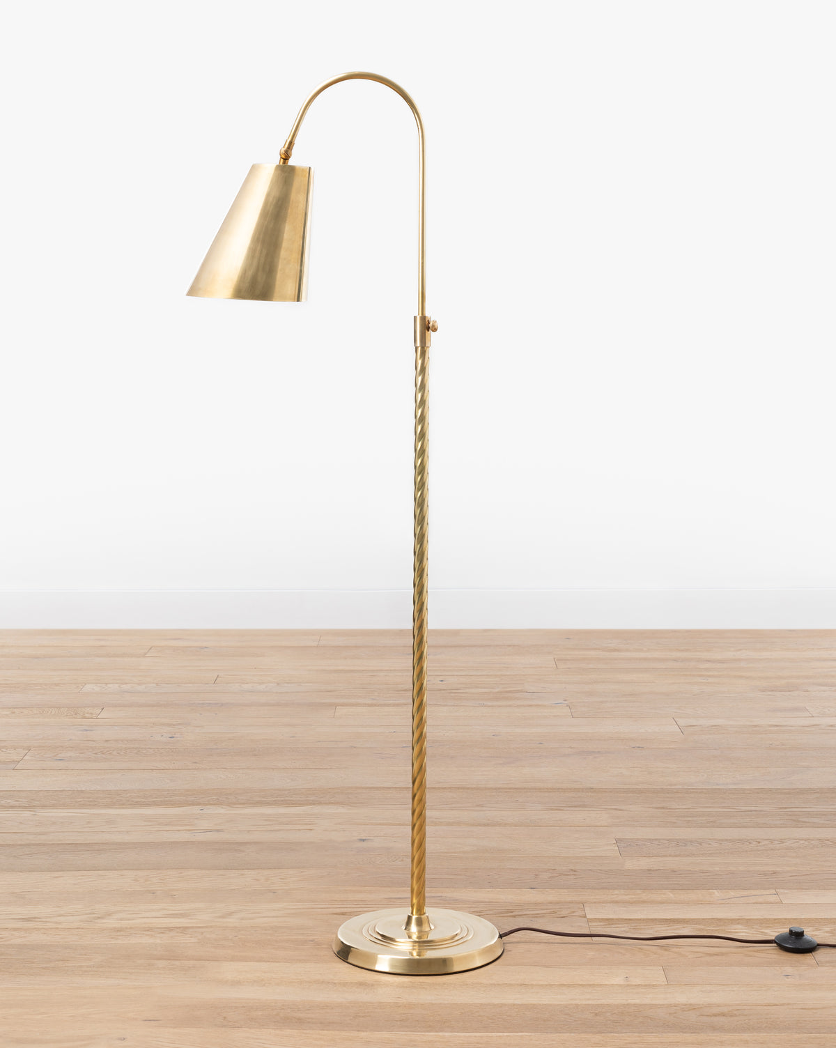 The Saylor Floor Lamp by Makerspalm features an elegant curved arm, conical shade, twisted brass stem, round base, and black power cord with switch—perfect for adding stylish lighting to your living room. McGee & Co.
