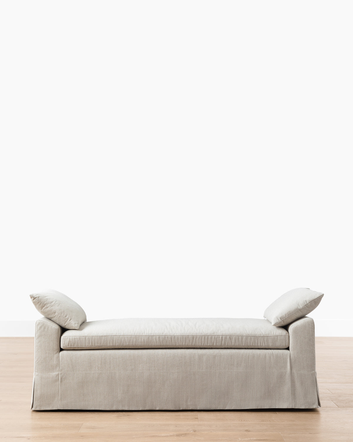The Makerspalm Scaletta Bench (Ready to Ship) features light gray linen upholstery and includes two matching angled pillows, shown on a wooden floor with a plain white background. McGee & Co.