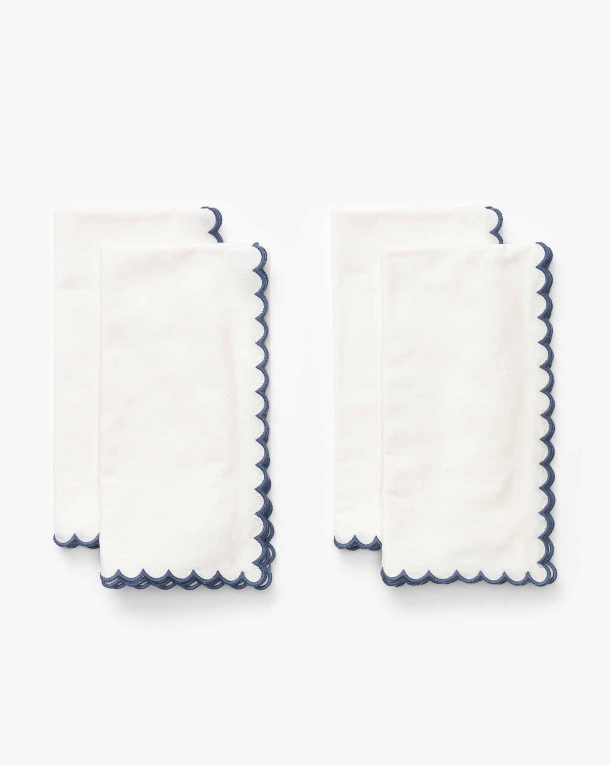 The Scallop Border Napkins (Set of 4) are neatly folded, each white cotton napkin featuring a scalloped navy blue trim—ideal for elevating any table setting, by McGee & Co.