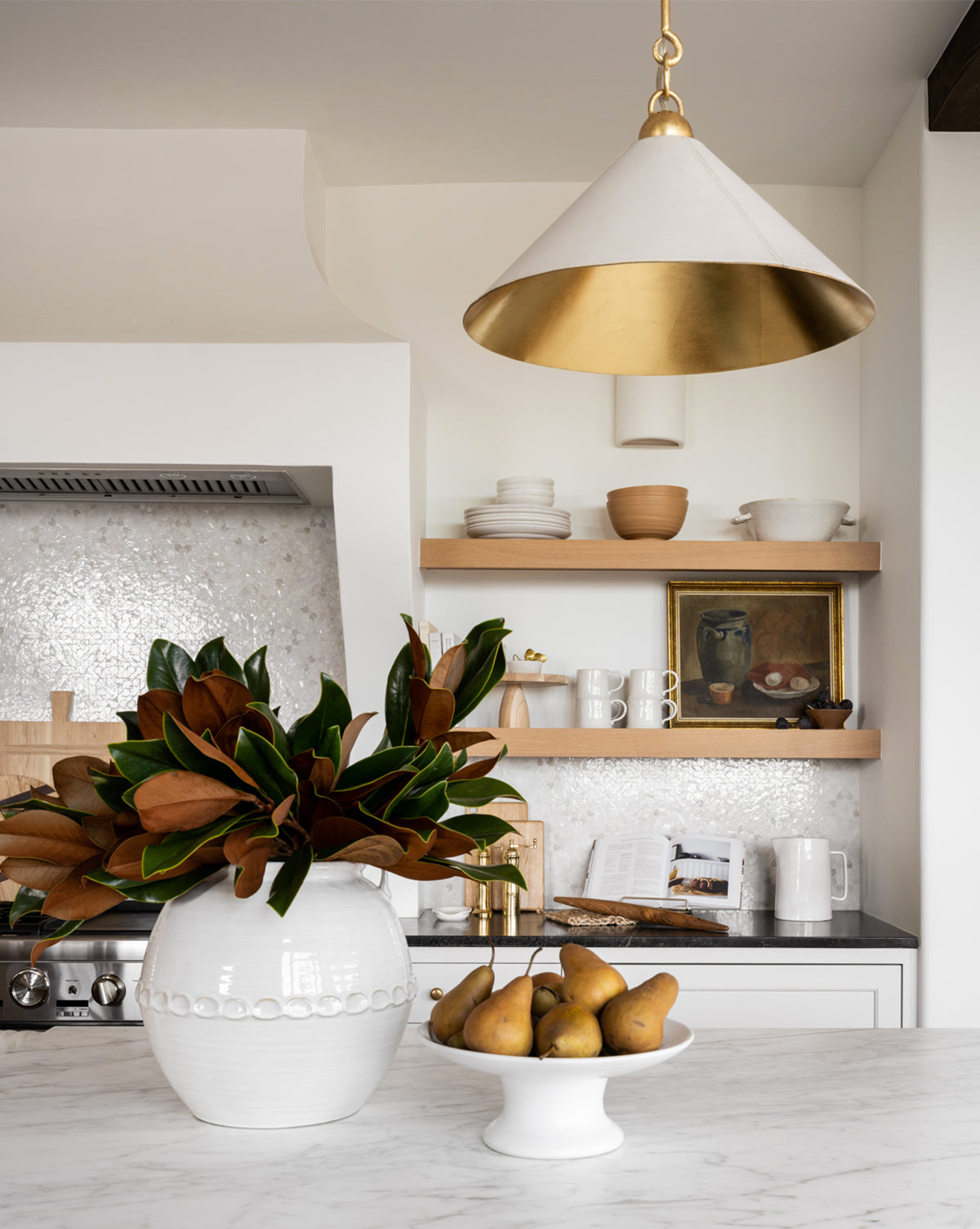 A modern kitchen features open wooden shelves with dishes and art, a white vase of magnolia leaves, a bowl of pears on marble, and elegant overhead lighting from the Scarlett Pendant, by McGee & Co.