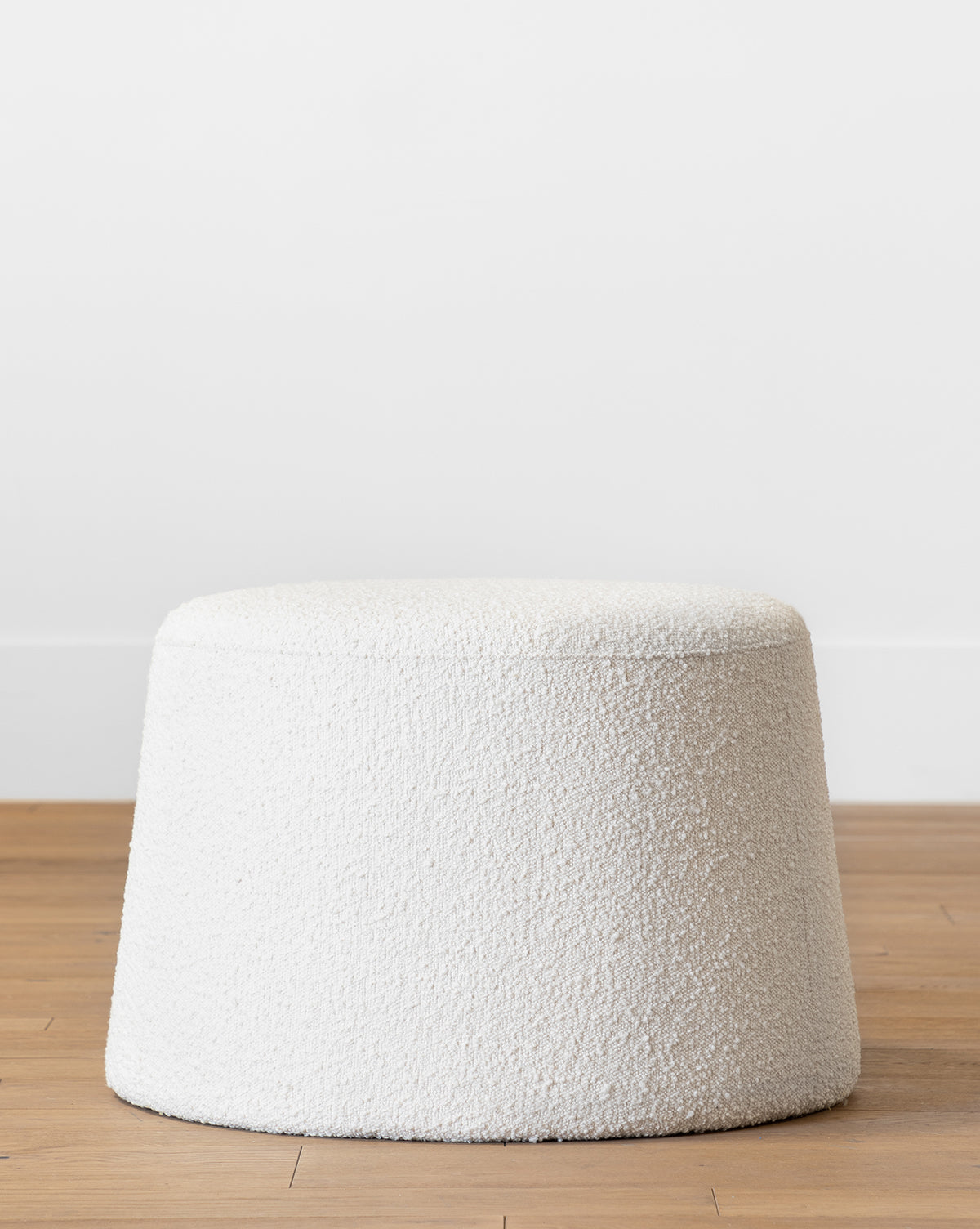 The Makerspalm Scout Ottoman features round, cream boucle upholstery with textured finish, resting on a light birch wood floor against a plain white wall by McGee & Co.