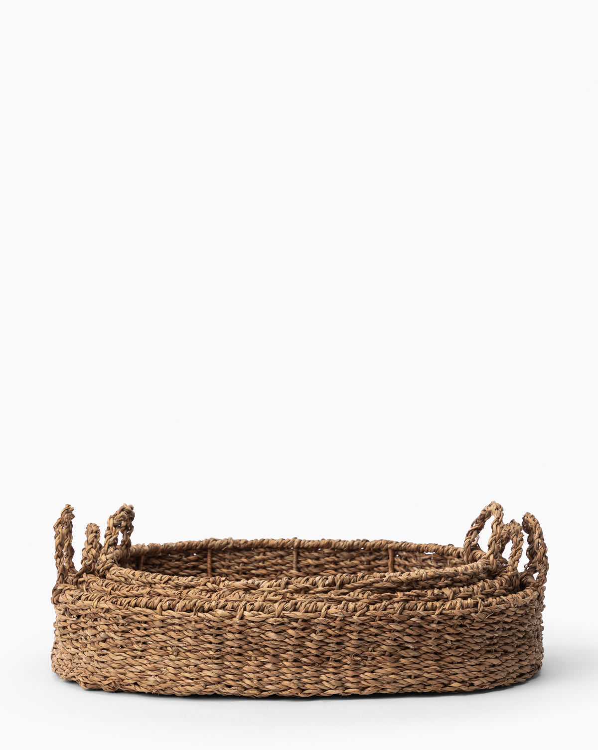 Three Seagrass Oval Trays with sturdy braided handles nest inside each other on a white background. Crafted from natural, textured seagrass, they combine beautiful details with stylish functionality as decorative trays.