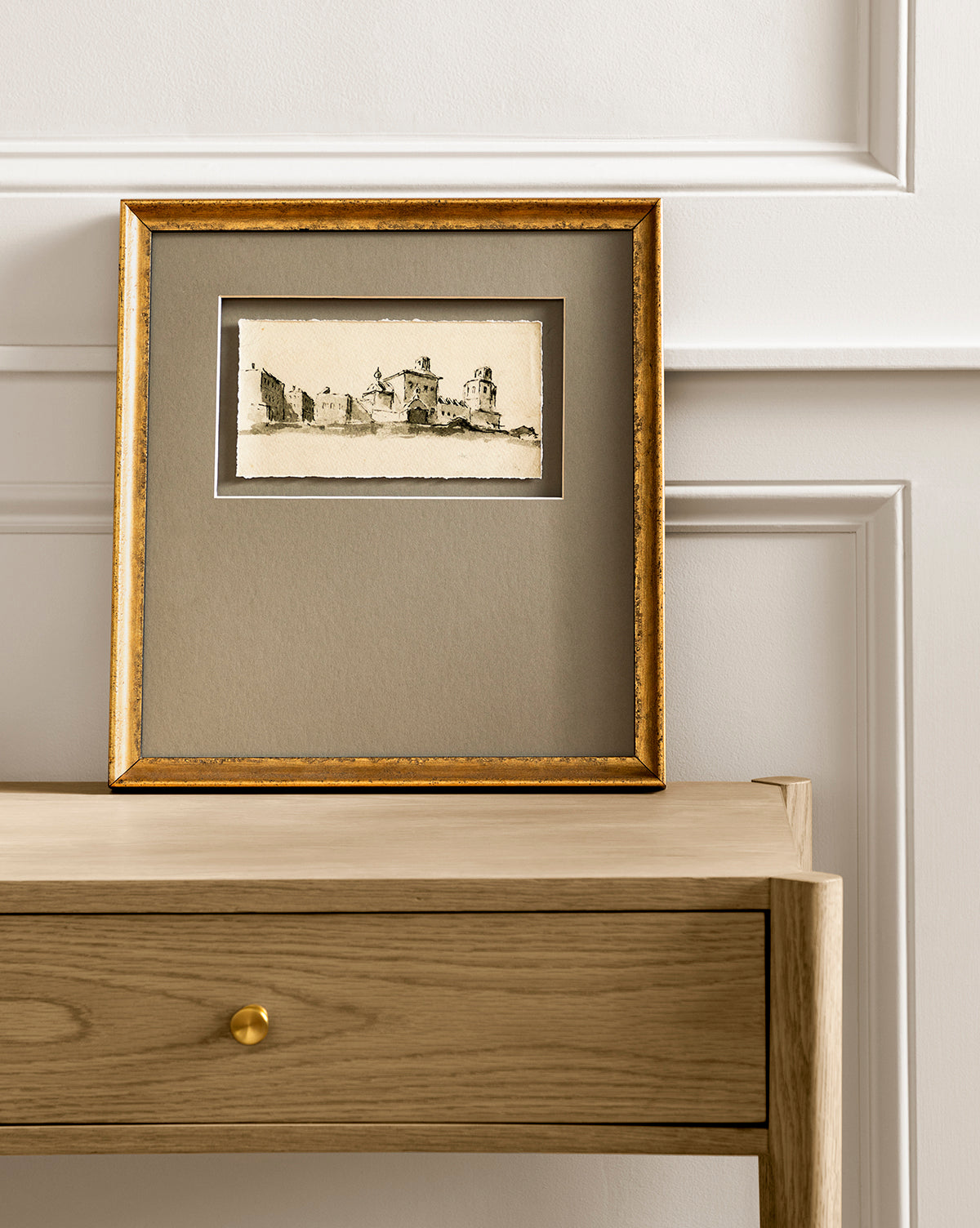 Sepia City, a framed black-and-white city sketch with a gold-edged frame and neutral mat, sits atop a light wooden drawer against a white paneled wall, creating an elegant, minimalist display.