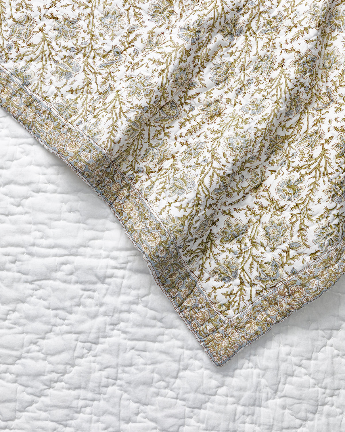 A close-up of the Seton Kids Quilt by Renuka Inc., featuring a leafy floral pattern in green and gold on white, draped over a plain white textured bedspread, McGee & Co.