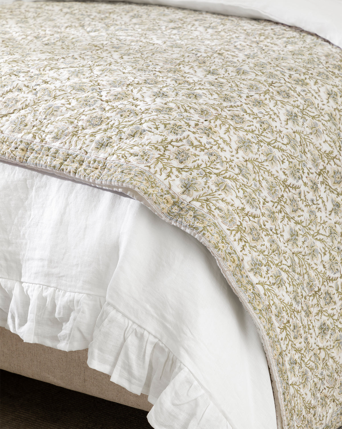 A close-up of a bed featuring the Seton Kids Quilt, showcasing its delicate green and beige floral motif and a white ruffled bed skirt draped gracefully over the edge, by McGee & Co.