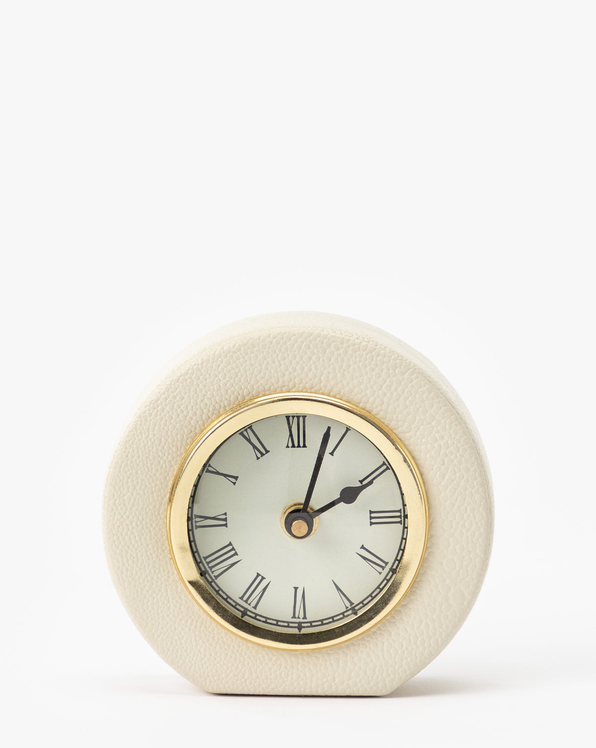 The Shagreen Table Clock features a round analog face with Roman numerals, gold trim, and sits in a textured cream shagreen leather case. Set to 2:05, this elegant clock is displayed against a plain white background.