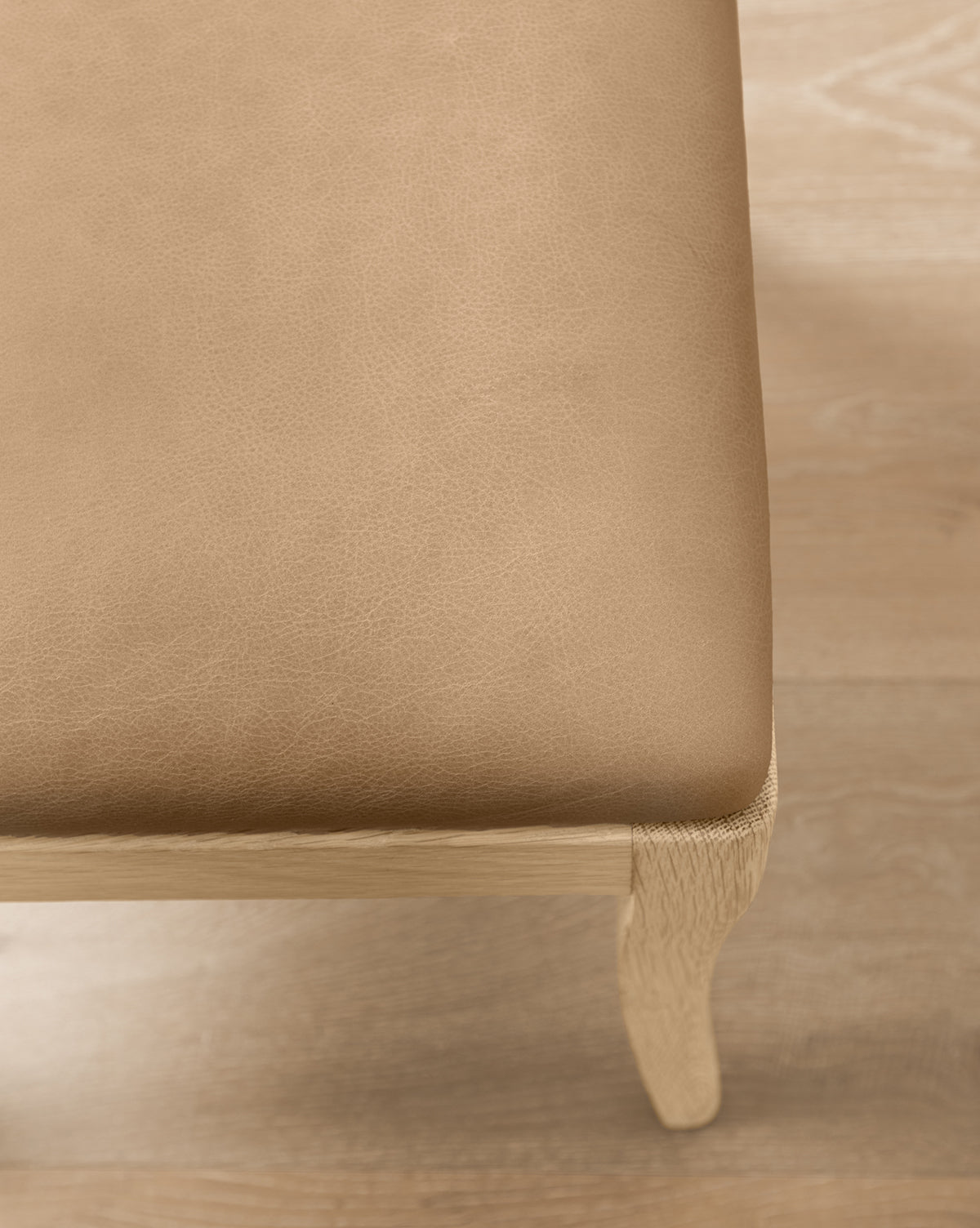 Close-up of the Shepherd Counter Stool by Makerspalm, featuring a light brown leather seat cushion with smooth performance upholstery and a curved white oak leg, all set on a light wood floor. McGee & Co.