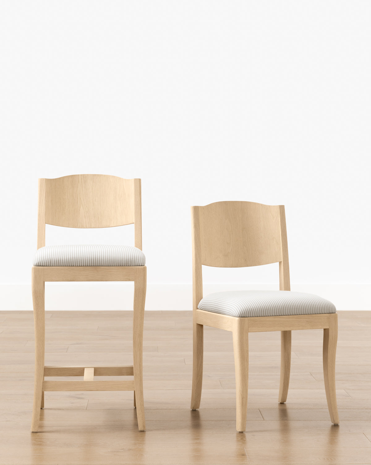 Two light wood chairs, including the Makerspalm Shepherd Counter Stool with a curved back and white-and-gray striped performance upholstery, sit side by side on a light wood floor against a crisp white wall. McGee & Co.