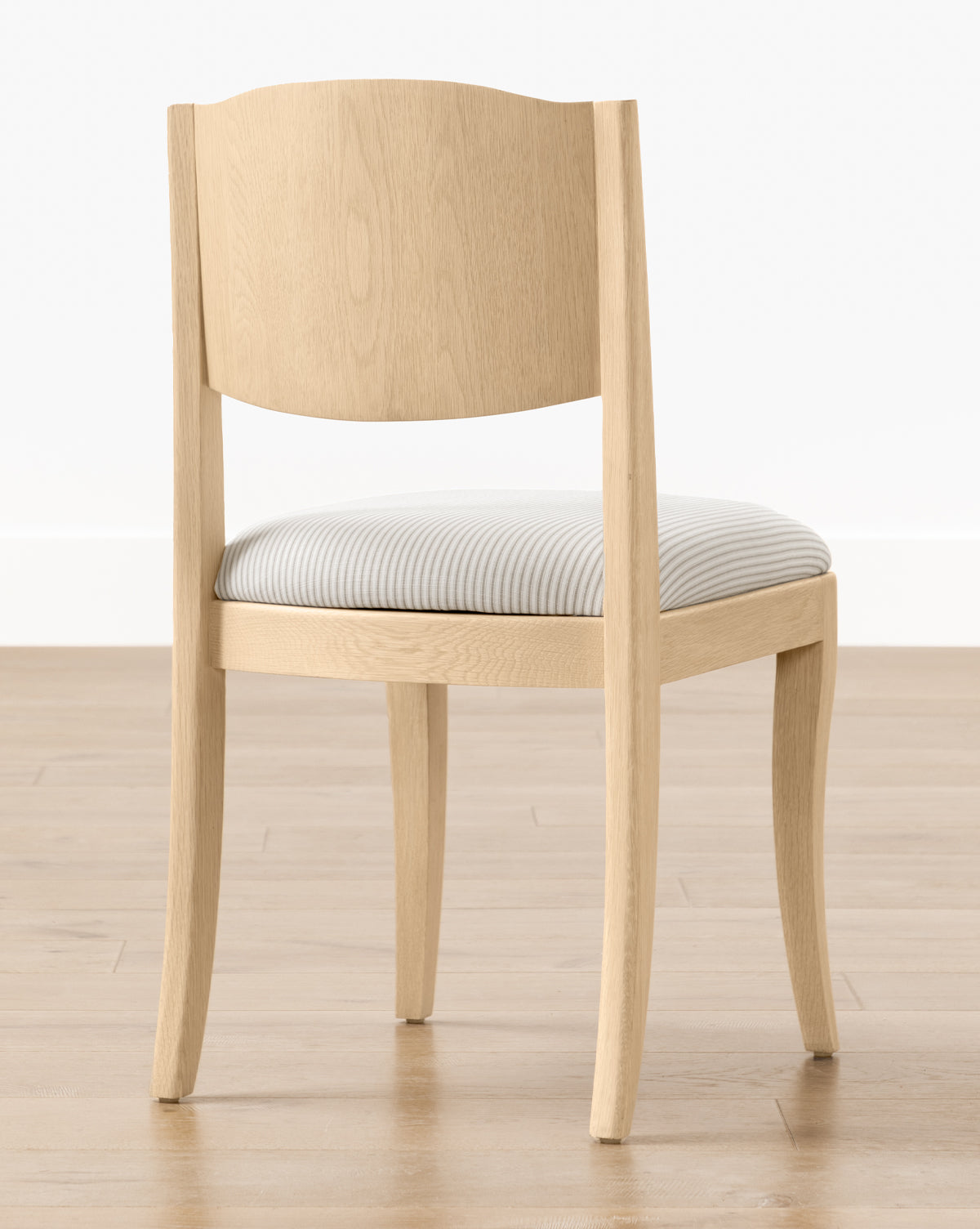 The Shepherd Dining Chair features light wood, a curved backrest, four legs, and a white and gray striped upholstered seat cushion. It is shown on a light wooden floor with a white wall background by McGee & Co.