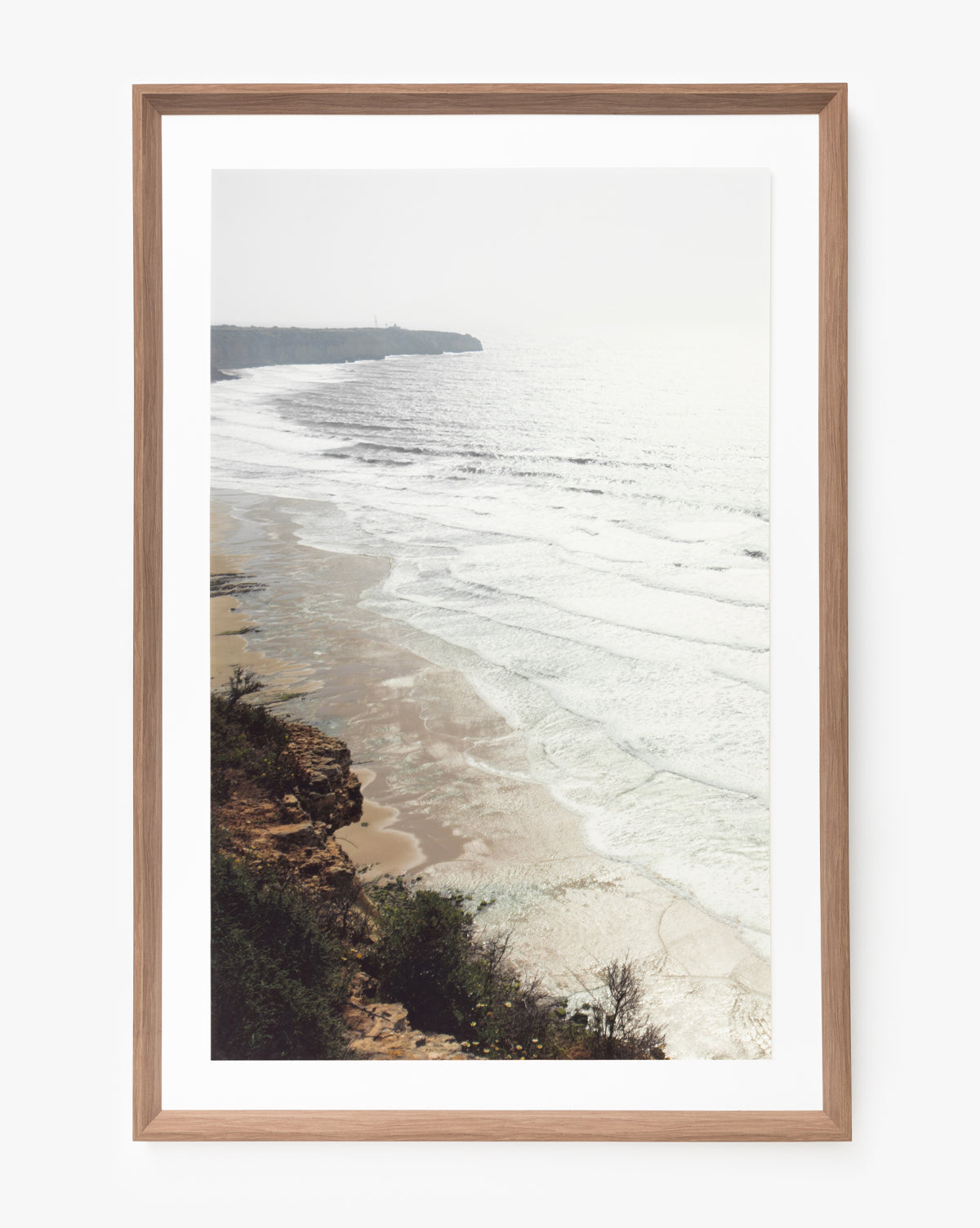 Shimmer of Wave is a giclee print featuring gentle waves on a sandy beach with rocky cliffs and sparse vegetation beneath a hazy sky—an ideal framed wall art piece in natural wood.