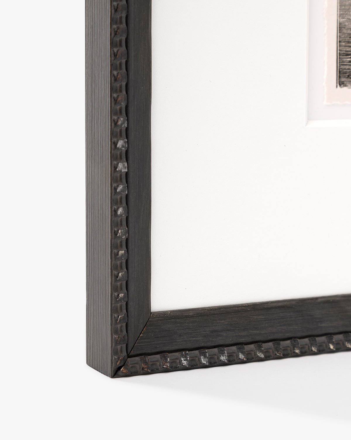 Close-up of the bottom left corner of a black, hammered metal-look frame with minimalist design and white mat board. The upper right reveals part of a seascape sketch, inspired by Shoals Engraving, by McGee & Co.