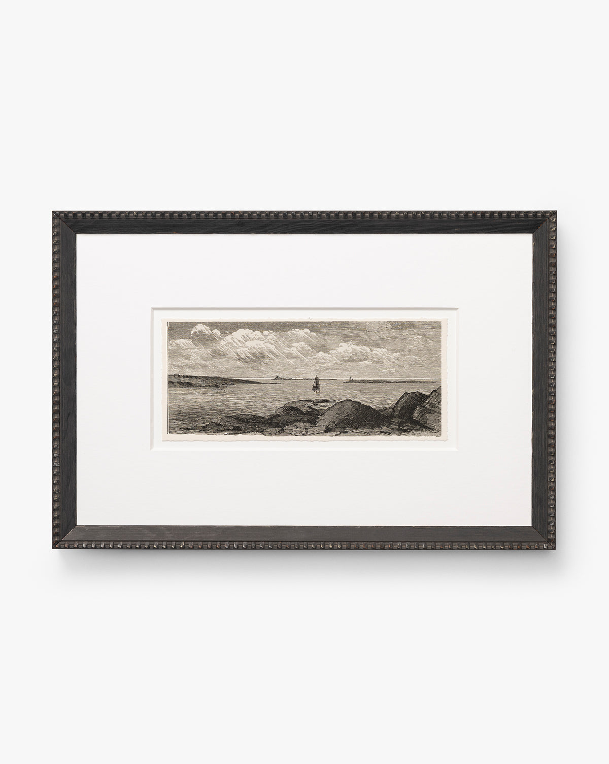The Shoals Engraving is a framed black-and-white seascape sketch featuring a sailboat on the horizon, cloudy sky, and rocky shore, centered in a wide white mat with an elegant black frame, by McGee & Co.