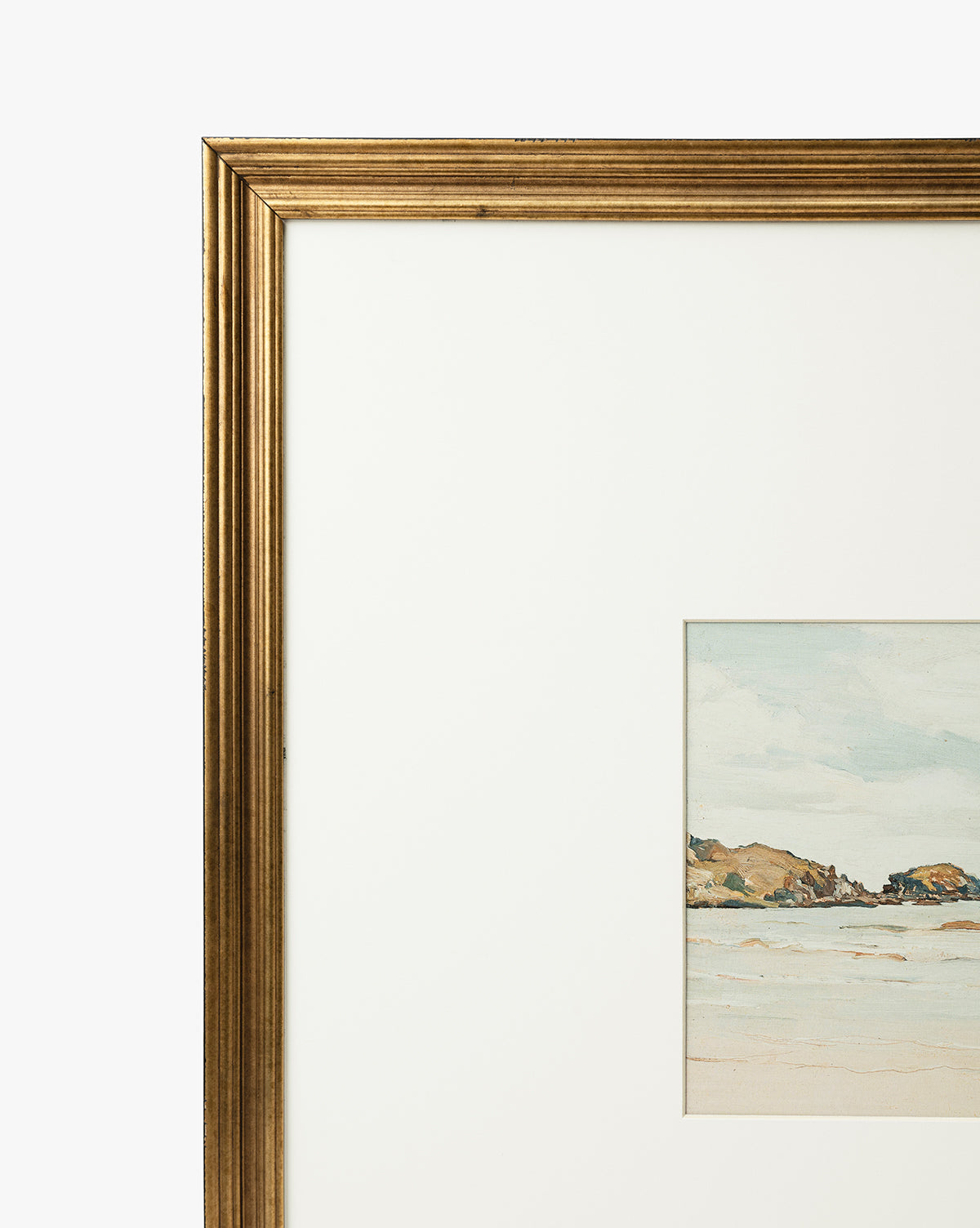 A close-up of the gold-framed Shoreline Rocks giclee print shows a coastal scene with shoreline rocks, sandy beach, and blue sky, set in a white mat. Only the left side of the frame and artwork are visible, by McGee & Co.