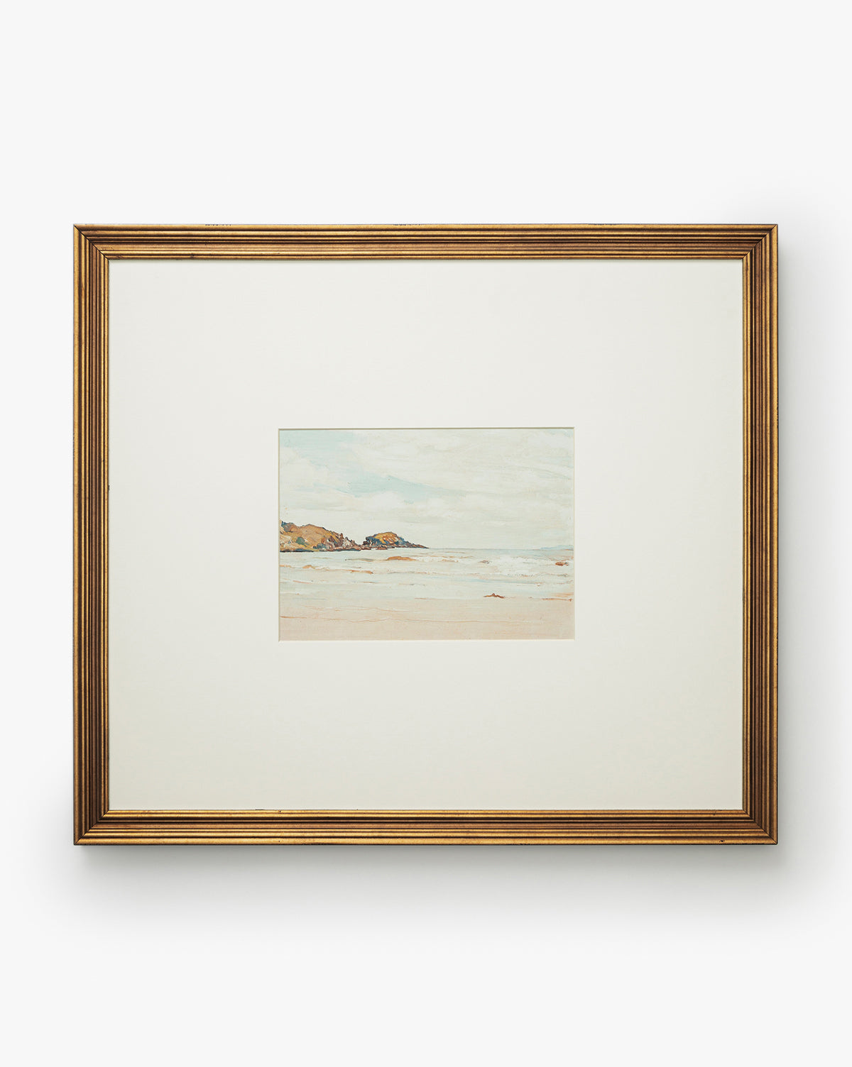 A framed giclee print titled Shoreline Rocks, featuring gentle waves and a sandy shore, is centered on a white mat within a gold frame, by McGee & Co.