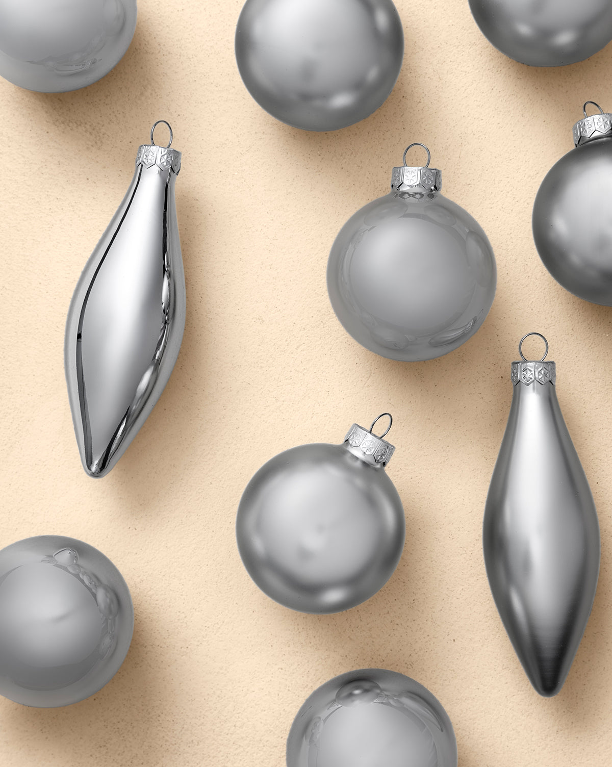 The Assorted Ornaments Silver (Set of 10) features shiny round and elongated ornaments with small top loops, arranged on a beige textured surface—ideal for enhancing your seasonal décor, by McGee & Co.
