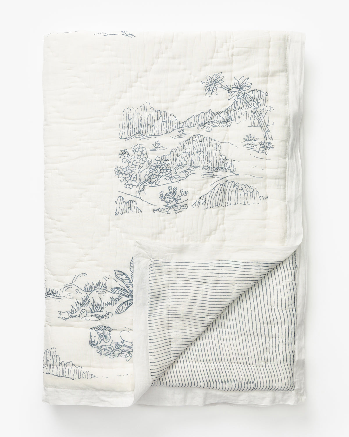 The Solano Block Print Kids Quilt by Indaba is a folded white cotton bedding quilt with blue botanical and landscape block print designs on one side and thin blue stripes on the reverse, shown on a plain white background. McGee & Co.