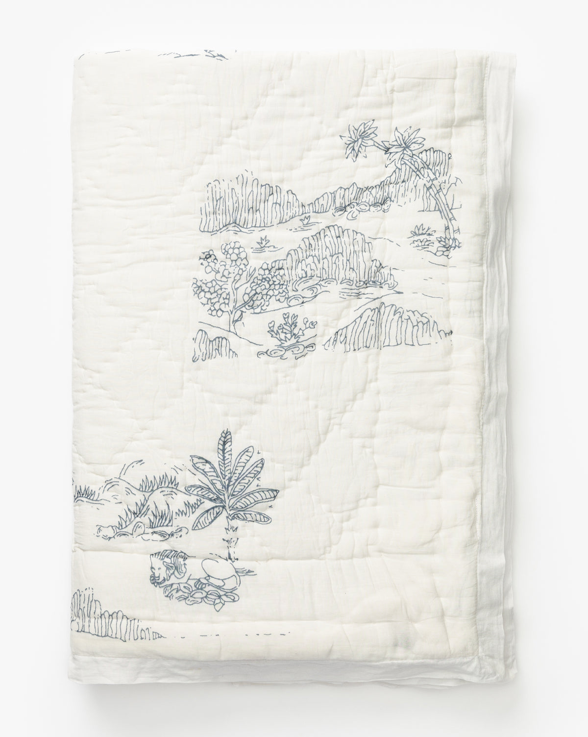 The Solano Block Print Kids Quilt by Indaba is a white cotton kids quilt featuring blue block print-style illustrations of landscapes with trees, hills, and a small dog, shown neatly folded against a plain white background. McGee & Co.