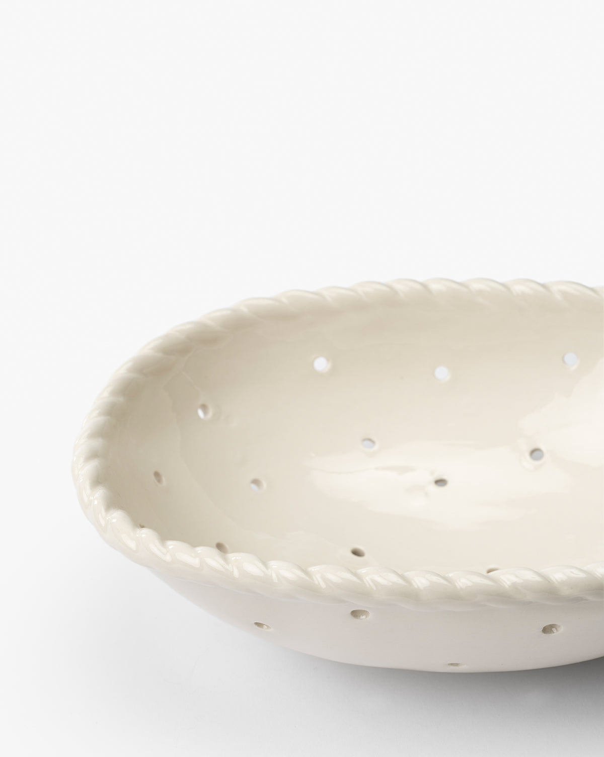 A close-up of the Solene Stoneware Bowl shows its white ceramic finish, braided rim, and evenly spaced holes. This elegant berry bowl enhances your kitchen decor, displayed against a plain white background.
