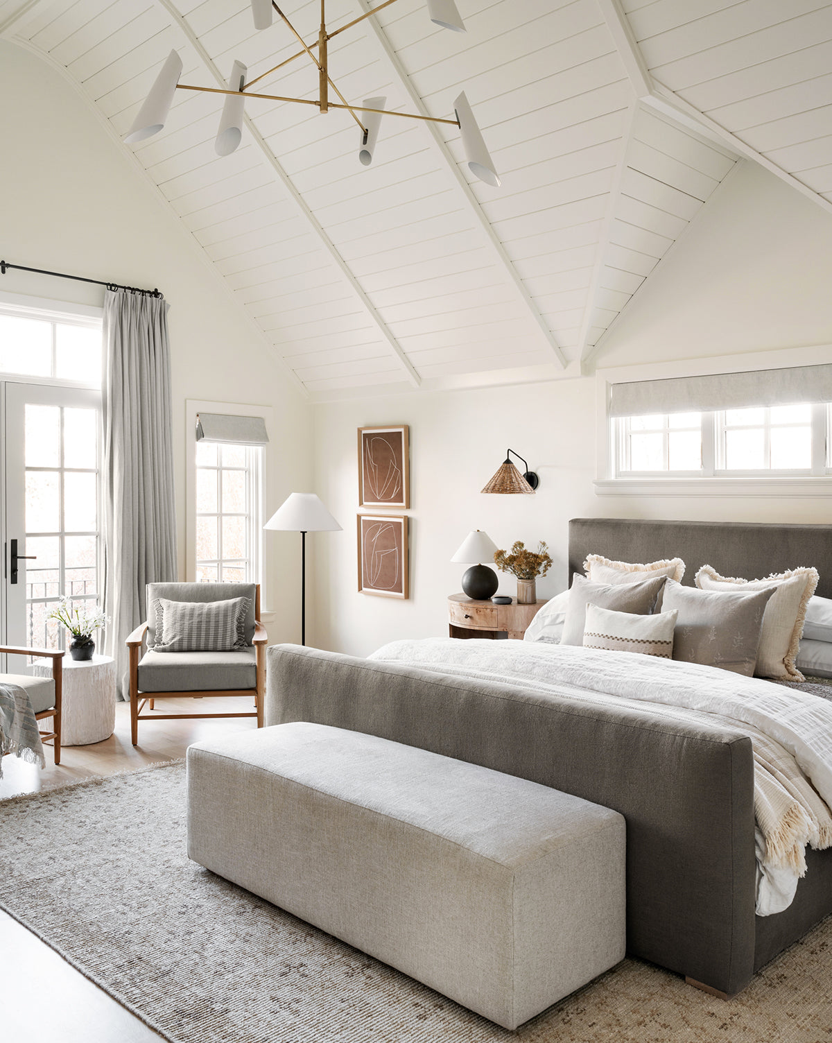 A modern, airy bedroom with vaulted white ceilings, neutral tones, a large bed with gray headboard, matching bench, armchair, wooden nightstand, soft lighting, wall art, and the Mali Hand-Knotted Rug enhancing natural light from windows and glass doors, by McGee & Co.