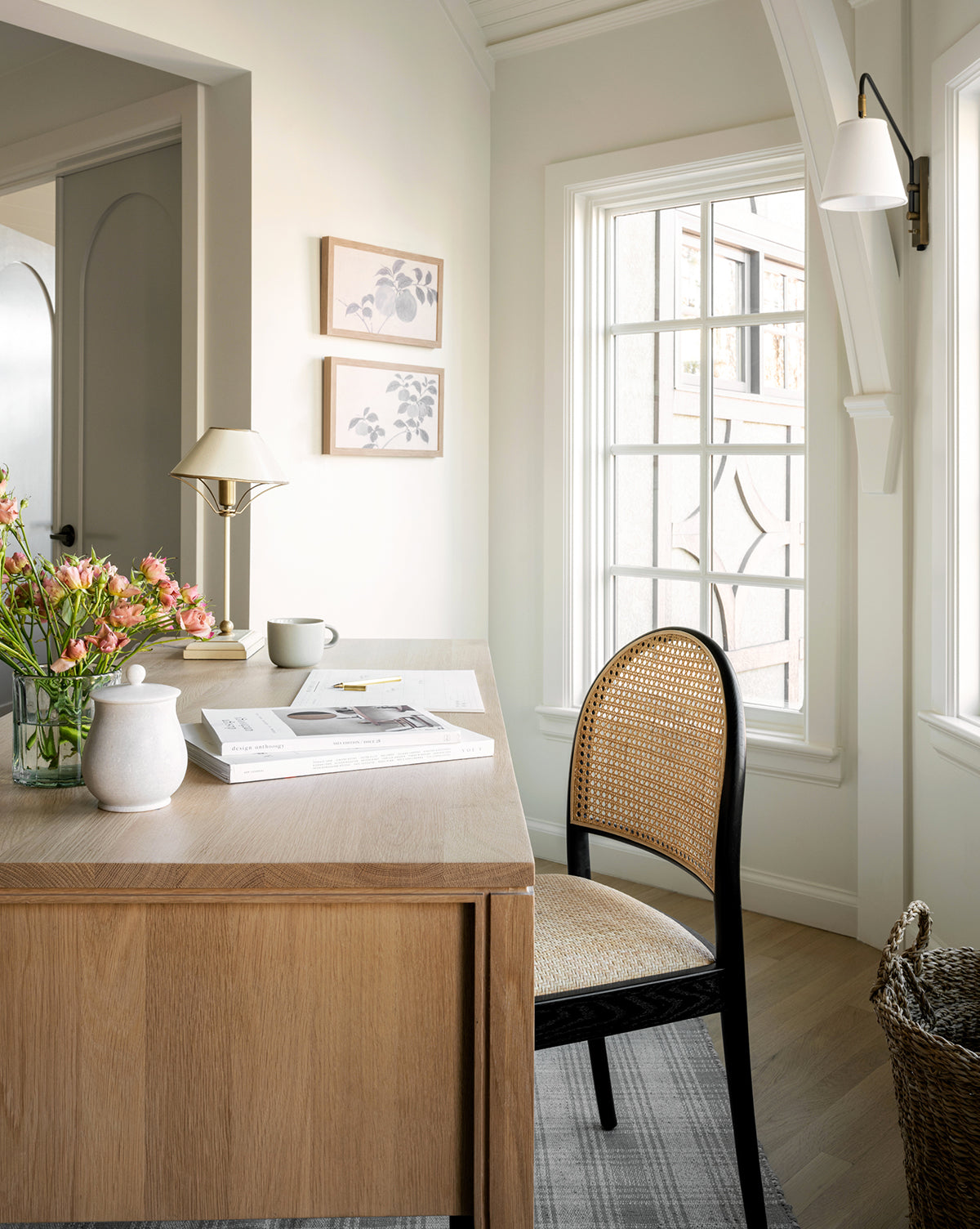 A bright, cozy home office features a wooden desk with books, a white vase, flowers, and a coffee cup. A woven chair sits by a sunny window, and the Inked Branches I giclee print on canvas hangs on the light-colored wall.